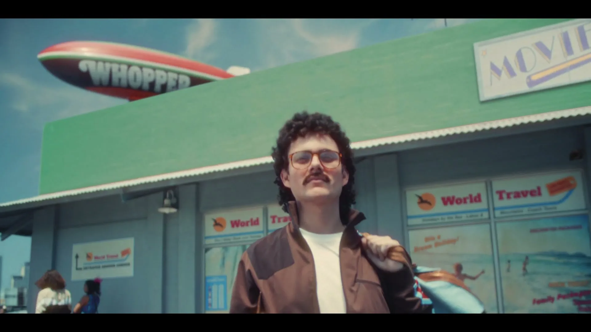 A man with a mustache and curly hair, wearing a brown jacket and glasses, stands in front of a travel agency with a "Whopper" blimp in the sky.