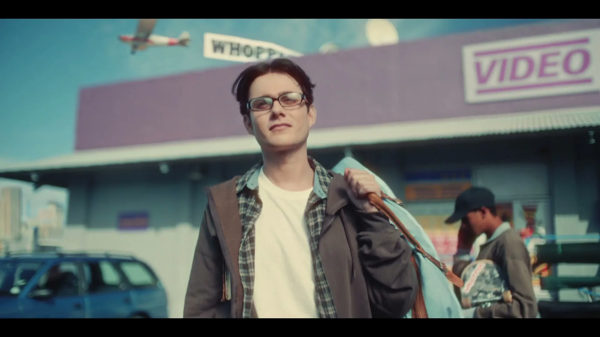 A young man with glasses and a plaid shirt stands outside a purple video store, looking up at a plane flying overhead with a "WHOPPER" banner.