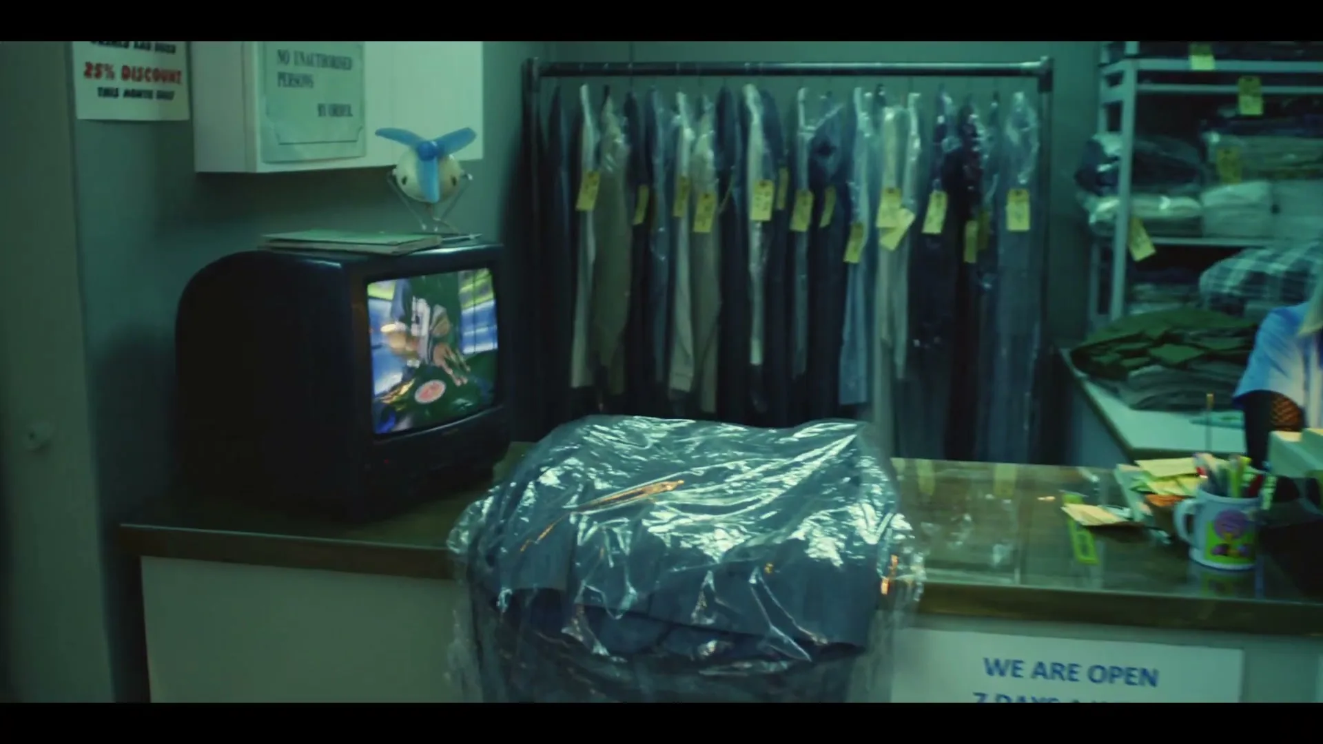 A young woman with blonde hair works at the counter of a laundromat, looking at a small TV playing a video of someone cutting a burger.