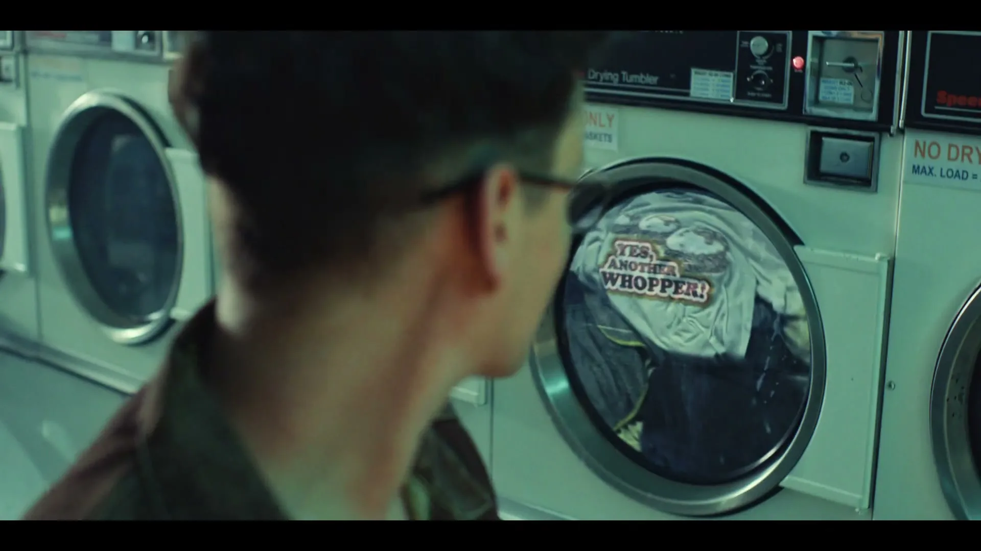 A young man with glasses and a goatee looks at a washing machine, where a t-shirt with "YES, ANOTHER WHOPPER!" is being washed in a laundromat.