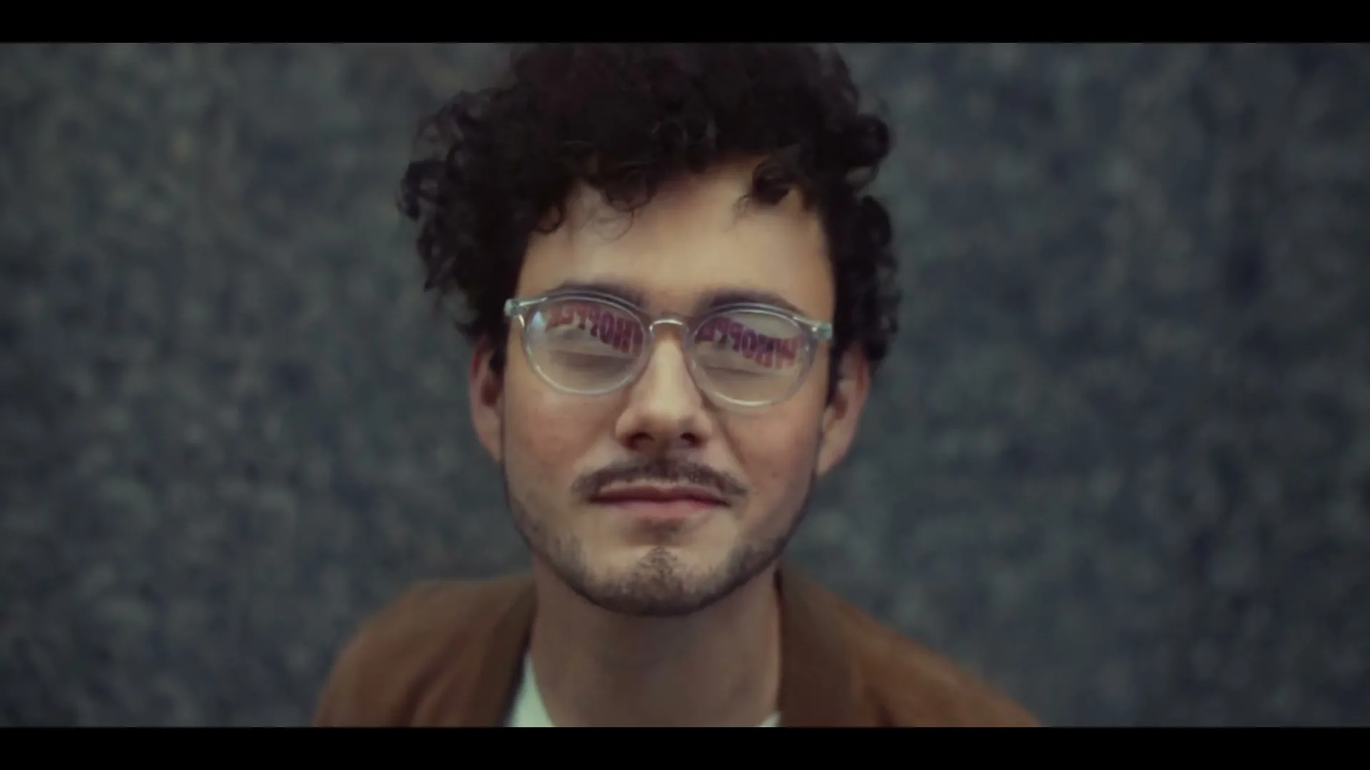 A young man with curly hair and glasses looks directly at the camera, with the word "WHOPPER" reflected in his lenses, against a grey wall.
