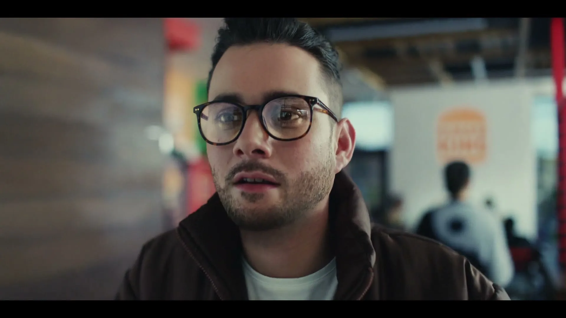 A young man with glasses and a brown jacket looks directly at the camera, then to the side, in a Burger King restaurant.
