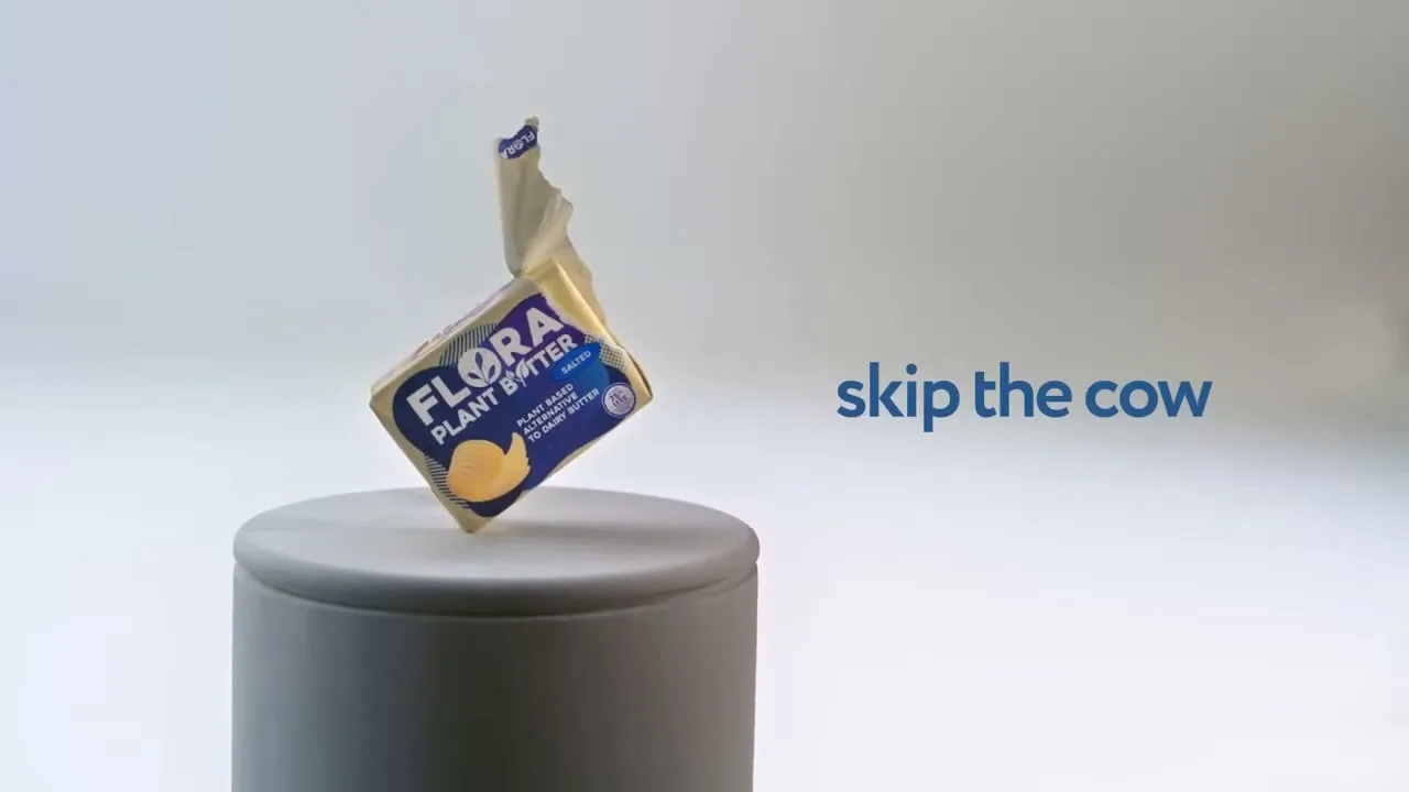 A block of Flora Plant Butter, partially unwrapped, balances on a grey cylindrical pedestal against a plain white background with blue text.