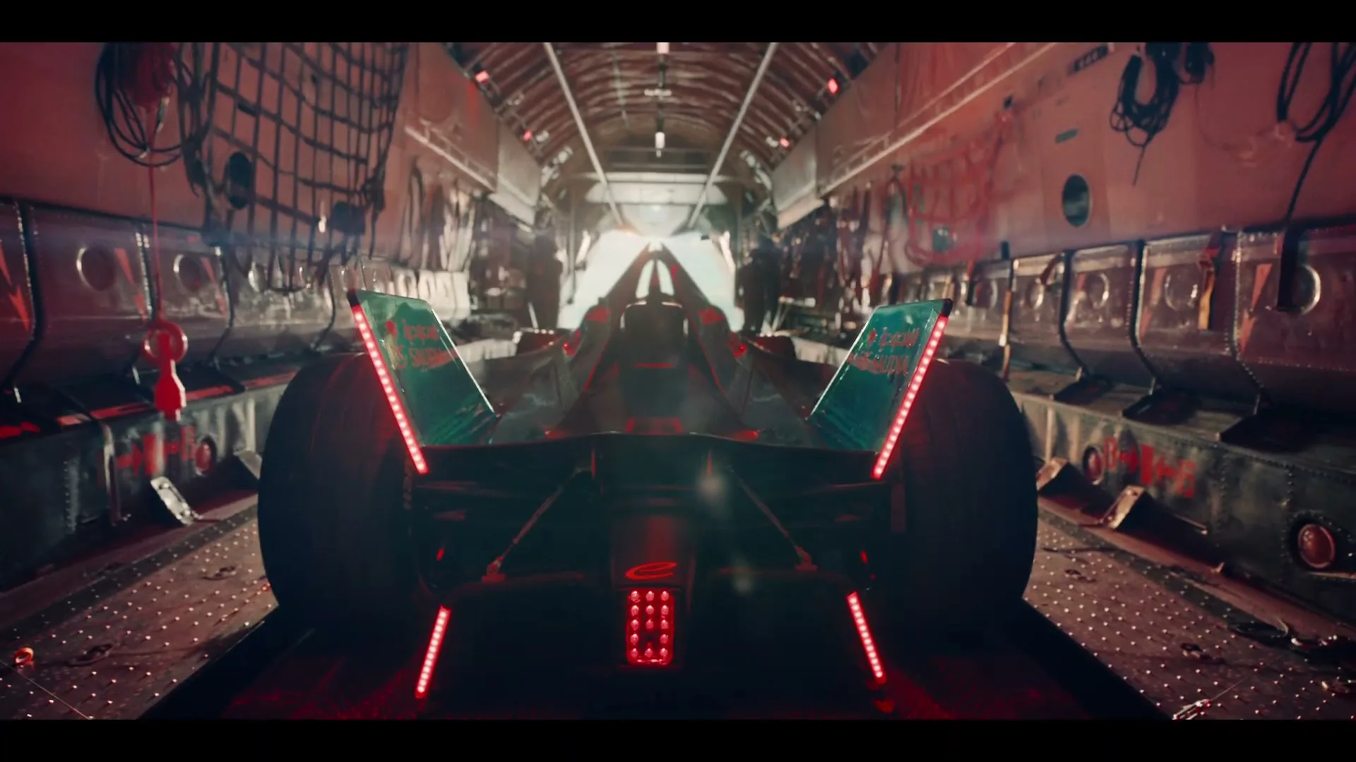 A Formula E race car, with glowing red lights, drives away from the camera down the cargo hold of a large transport plane.