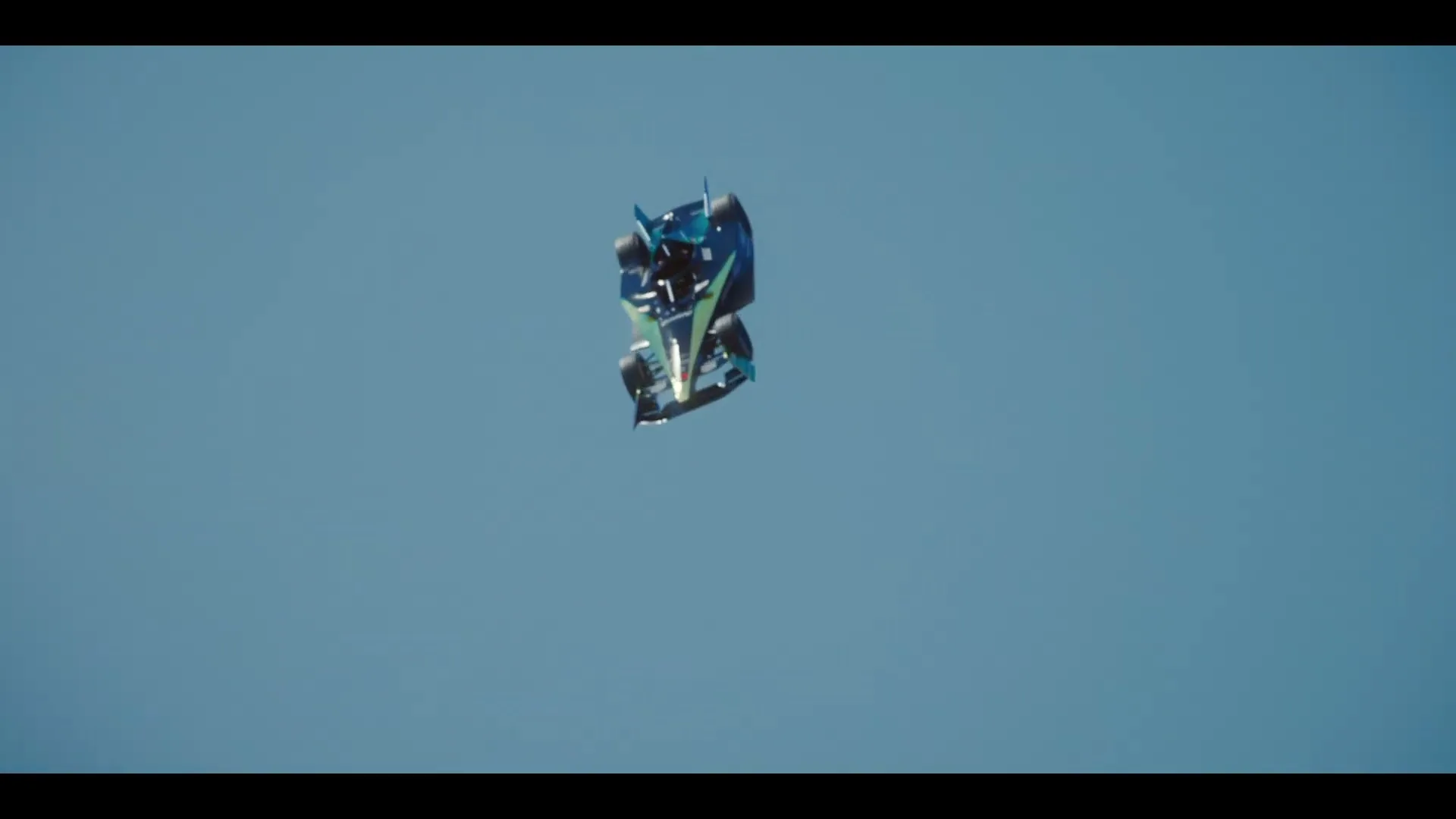 A Formula E race car, blue and black, flips through the air against a clear blue sky, showcasing its underside.