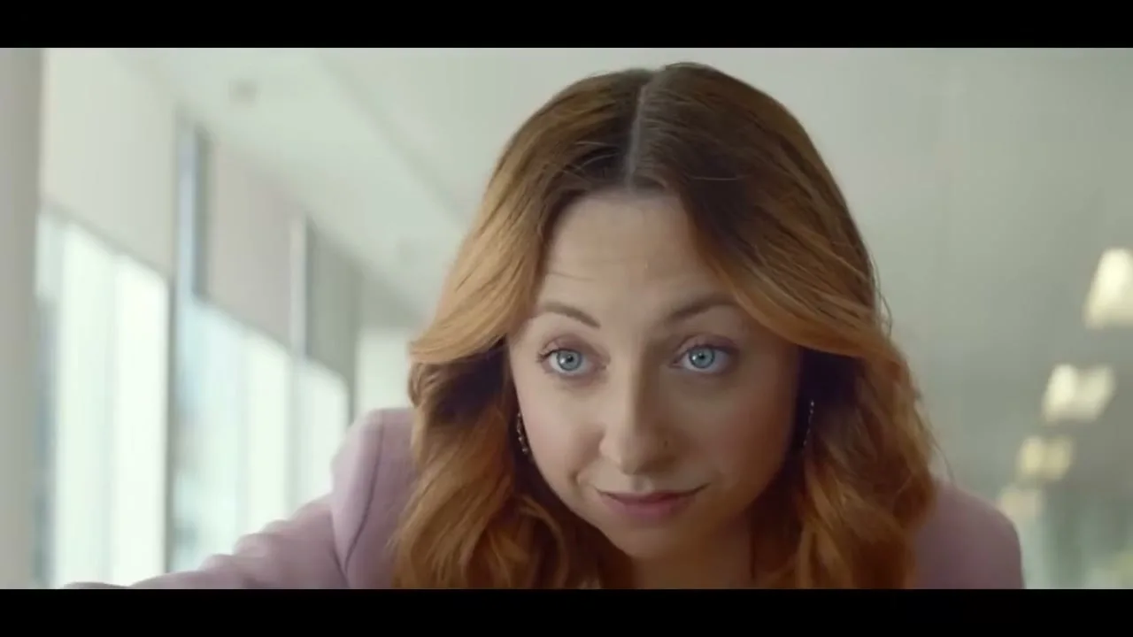 A woman with red hair and a pink blazer looks up with wide blue eyes and a slight smile in an office setting.