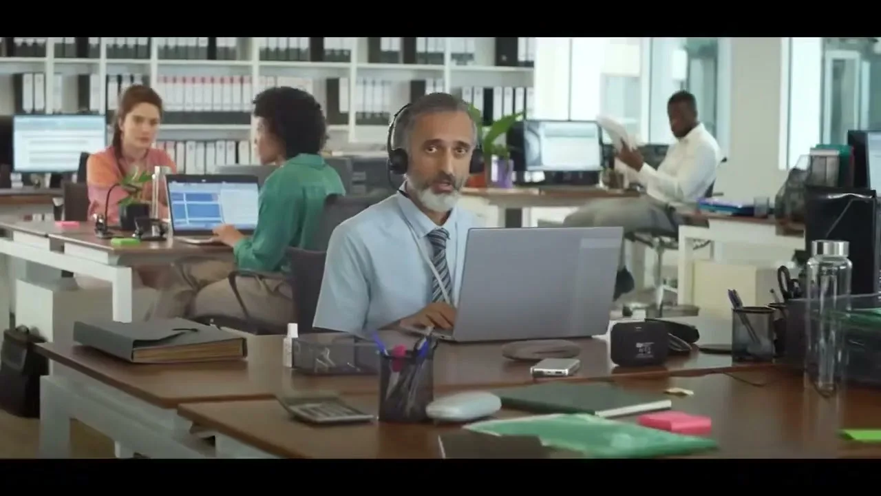 A middle-aged man with a headset works on a laptop at his desk in a busy open-plan office, looking directly at the camera with a neutral expression.