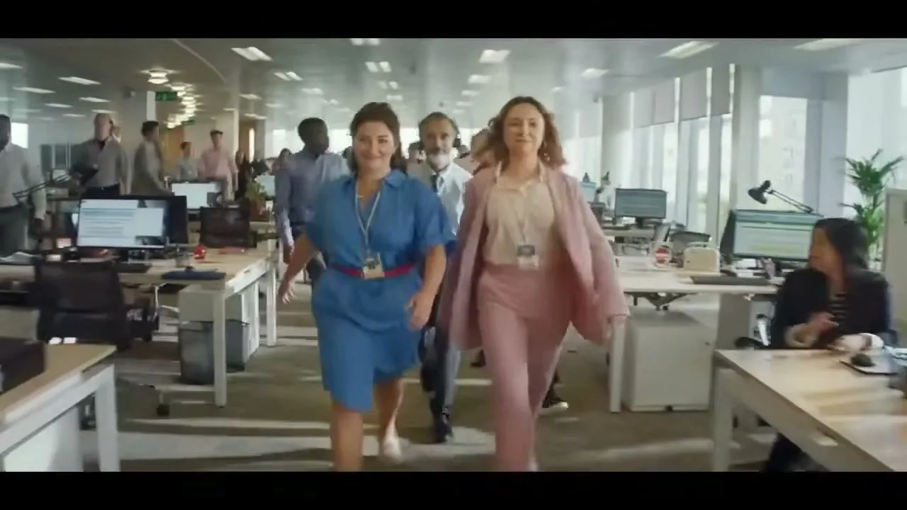Two women in an office, one in a blue dress and one in a pink suit, walk towards the camera, raising their eyebrows and smiling, as other office workers do the same in the background.