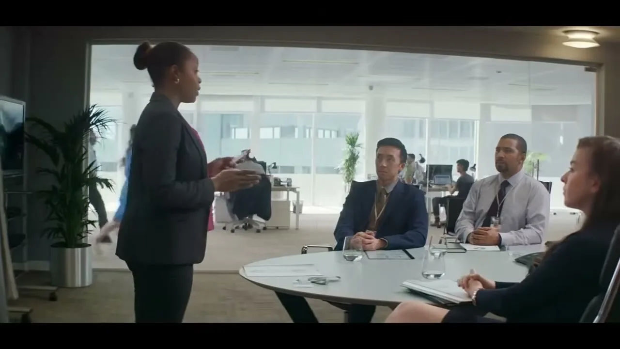 A Black woman in a business suit stands and speaks to three colleagues seated around a conference table in a modern office.