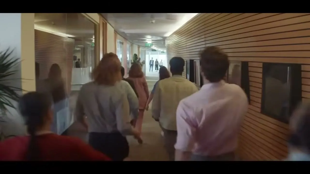 A diverse group of office workers, seen from behind, rush down a modern office hallway with wood-paneled walls and glass offices.