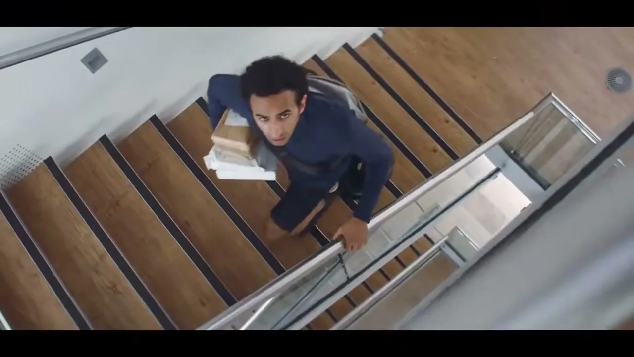 A young man with dark curly hair, wearing a blue long-sleeved shirt and carrying a stack of papers, climbs a modern wooden staircase, looking up with a surprised expression.