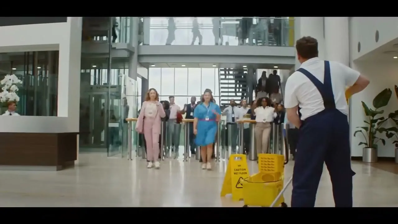 A male janitor mops the floor in a modern office lobby as a diverse group of office workers walk past him, raising their eyebrows.