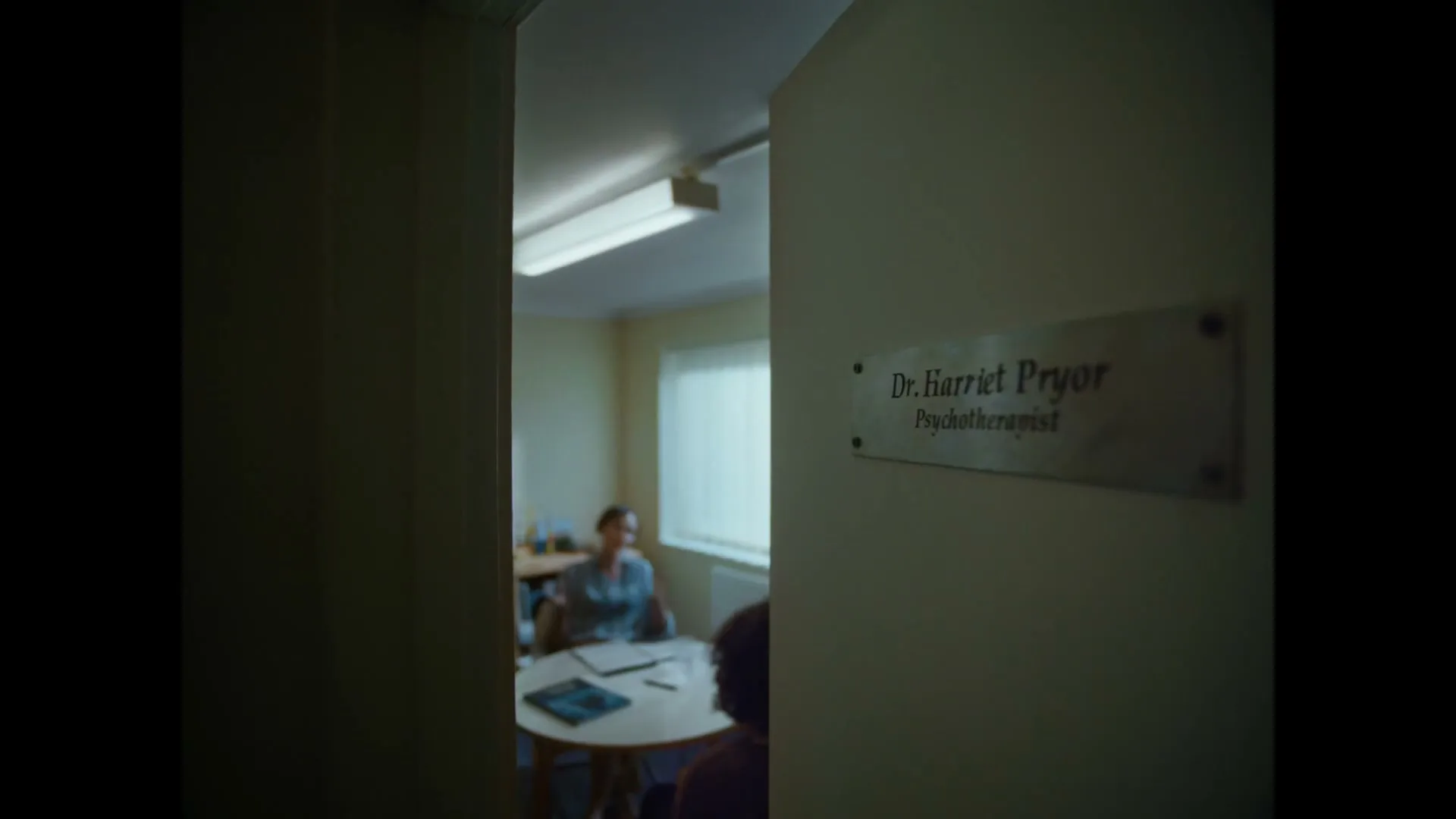A slightly ajar door reveals a blurred view of a psychotherapist and a patient sitting at a table in a brightly lit office.