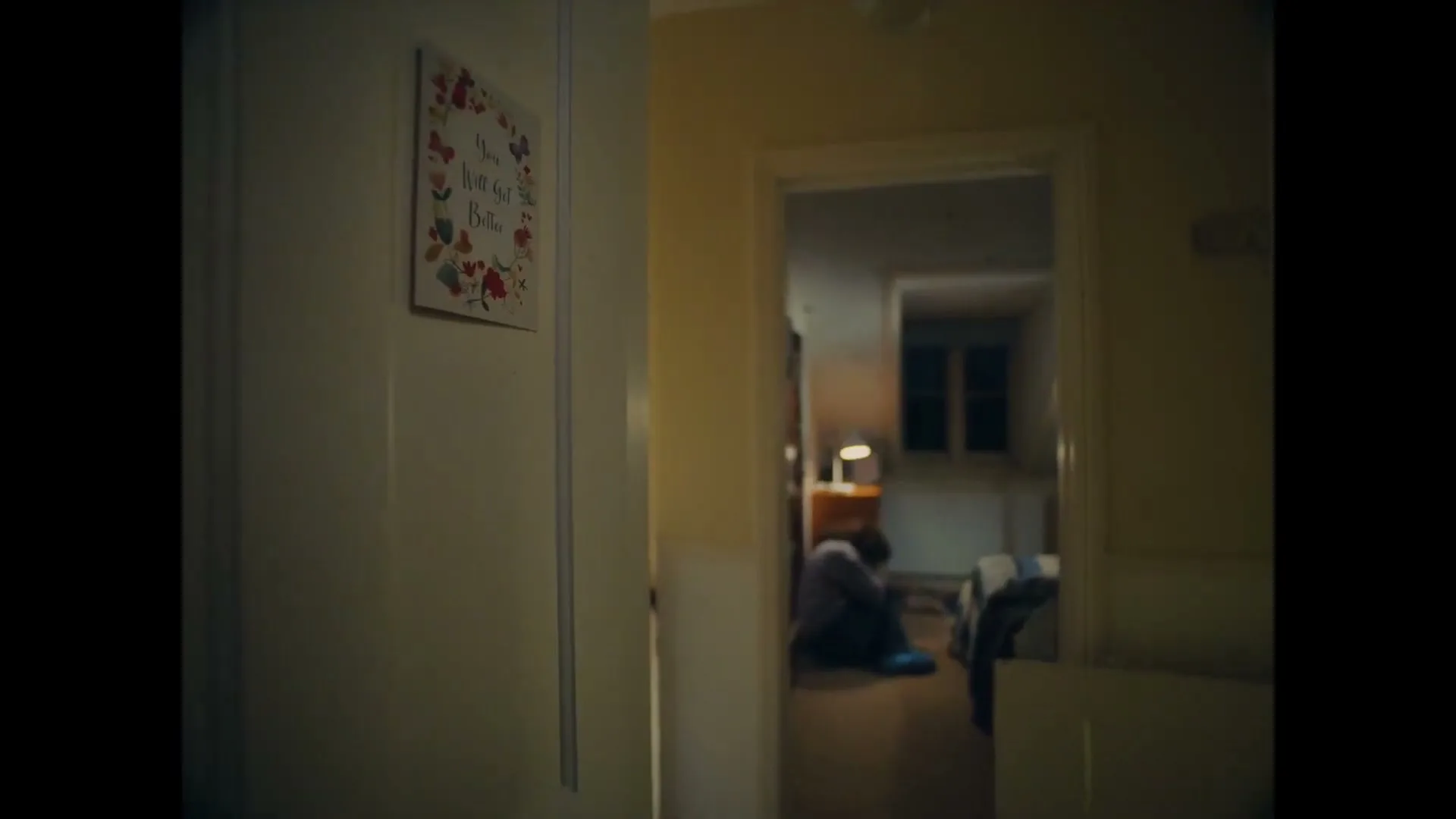 A person sits on the floor in a dimly lit room, seen through a doorway, with a "You Will Get Better" sign on a wall in the foreground.