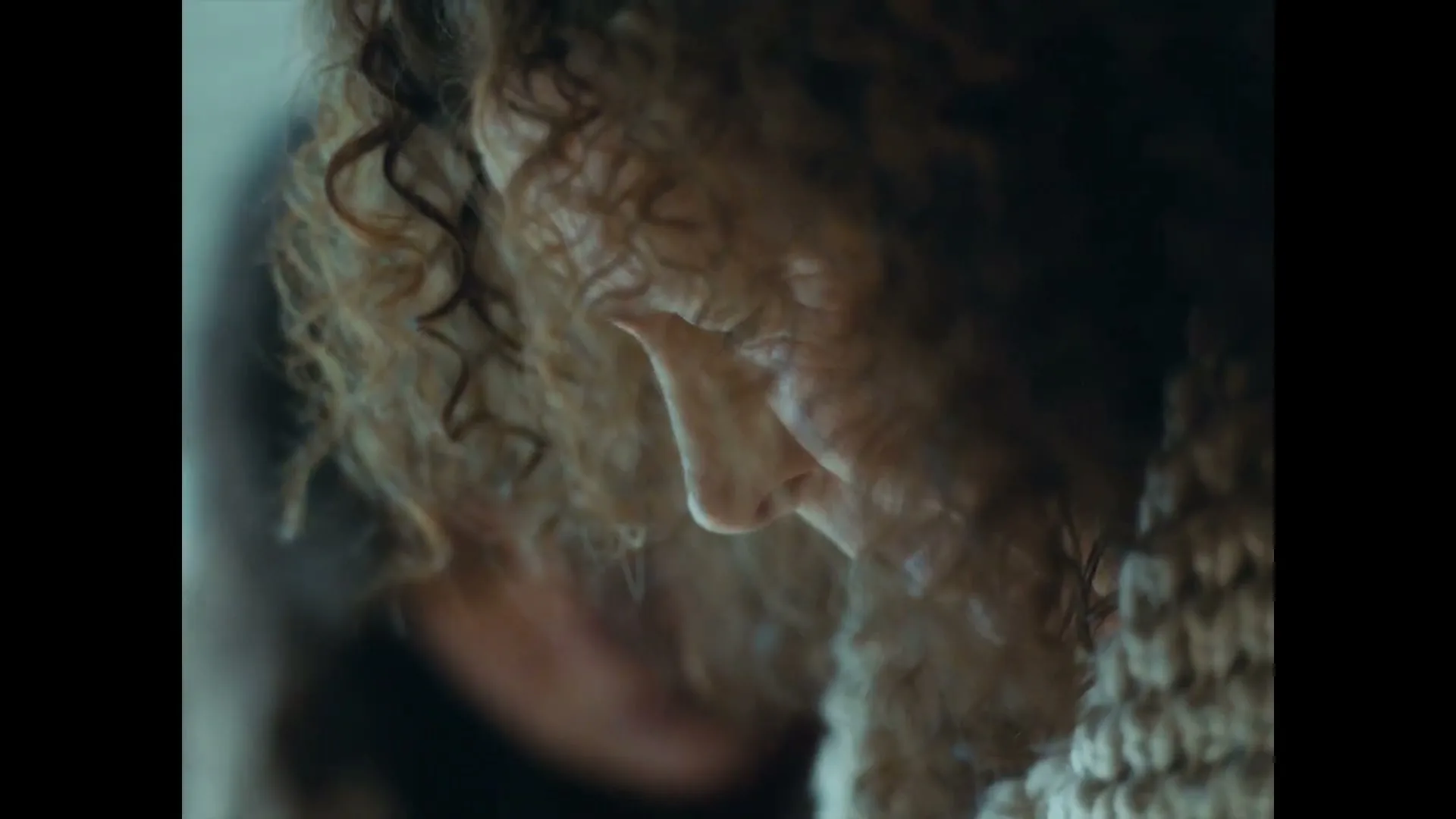 A person with curly hair and a knitted garment looks down, appearing distressed in a dimly lit interior.