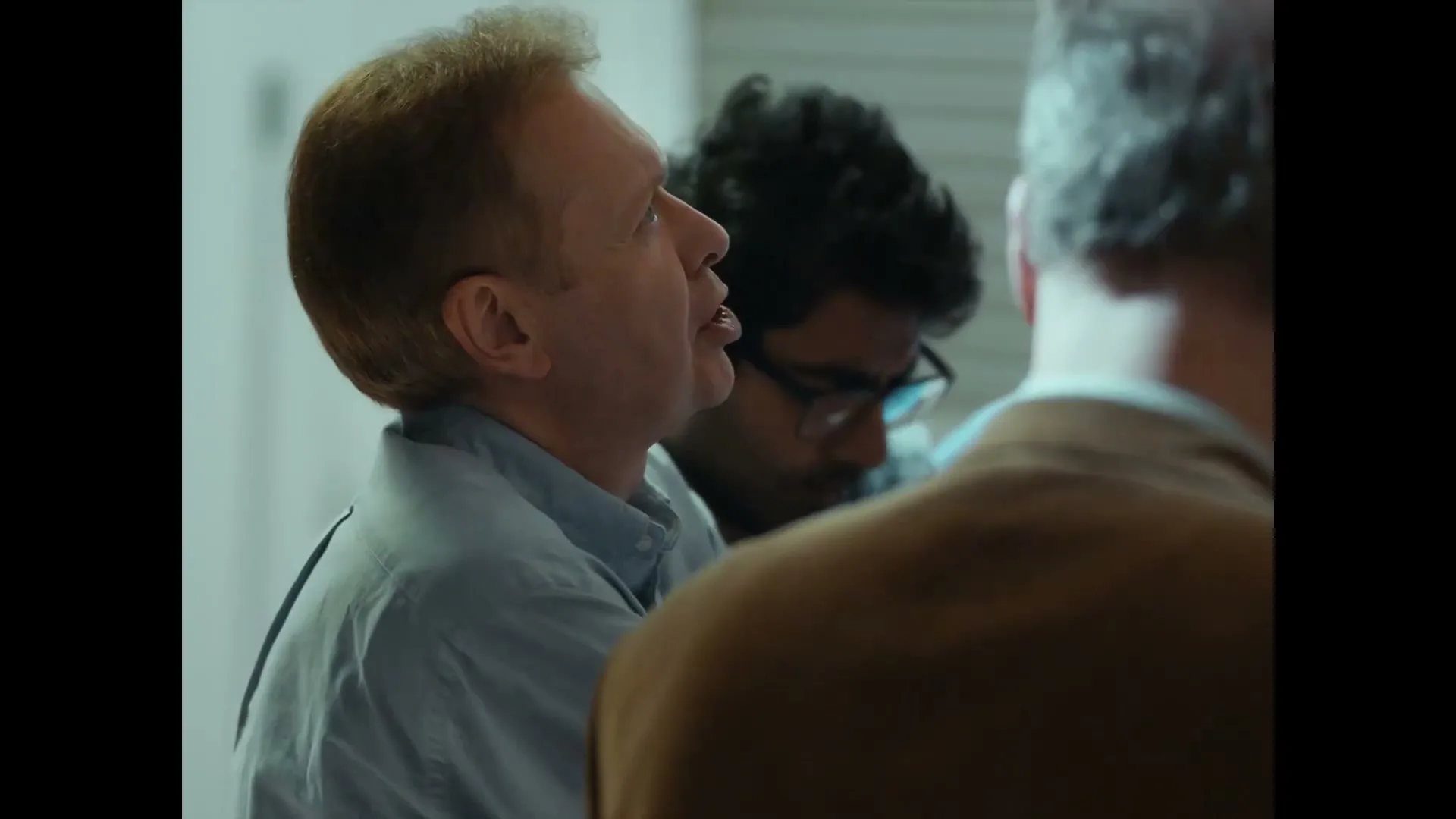 A man with reddish-blonde hair and a light blue shirt looks up and to the right, while two other men are blurred in the background.