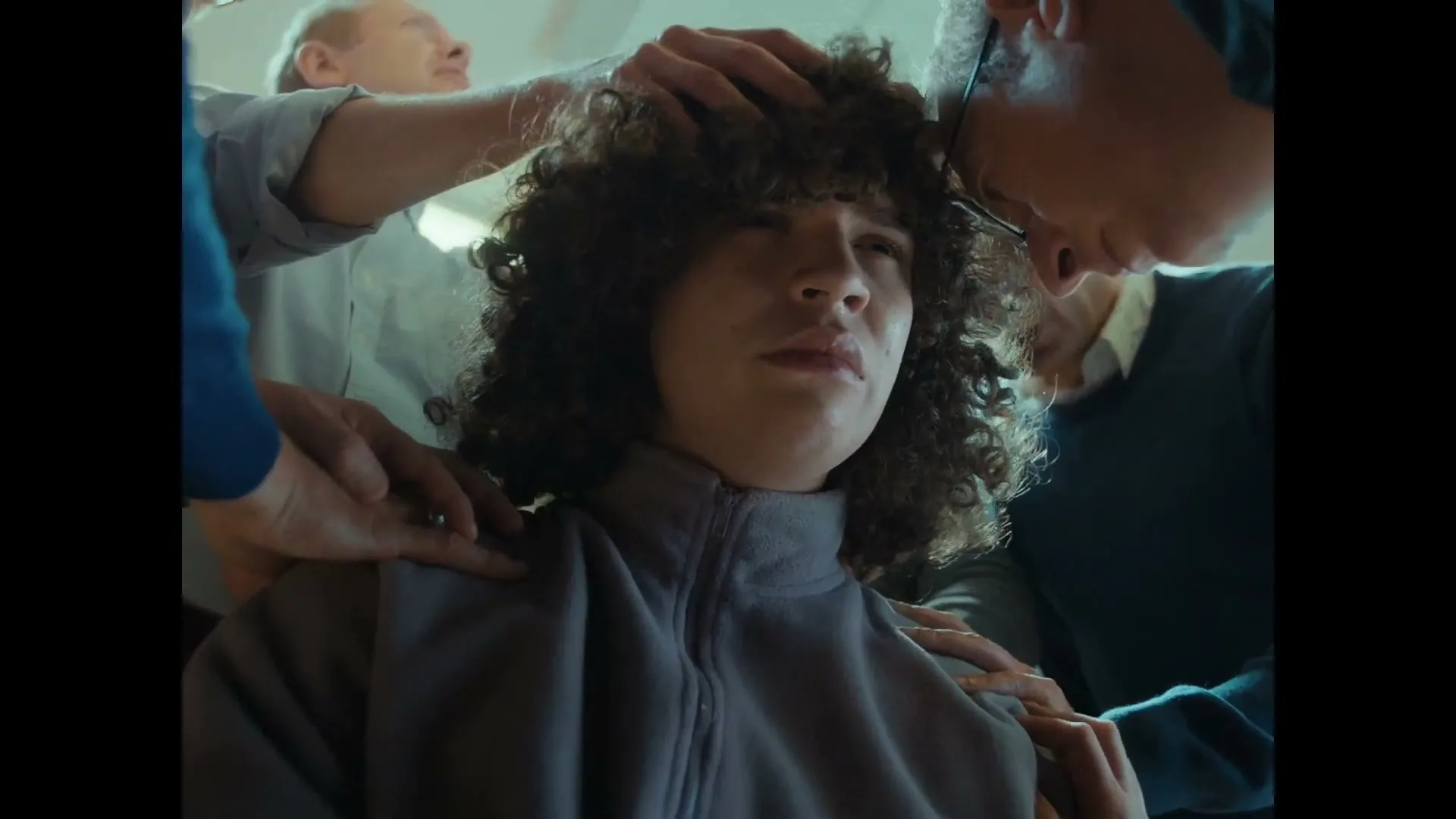 A young person with curly hair sits surrounded by adults touching their head and shoulders, looking distressed in an interior setting.