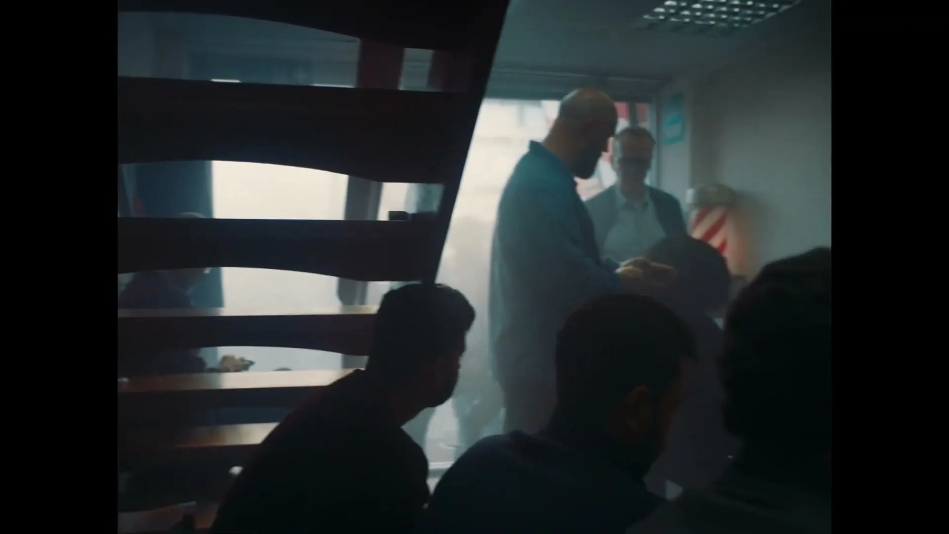 A camera pans right from a wall of mugshots to a barbershop, where a barber shaves a man's head while other men wait.