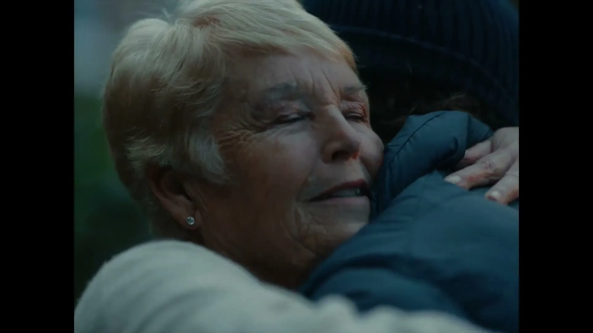 An older woman with short blonde hair embraces a younger person, her eyes closed and a look of relief on her face, outdoors.