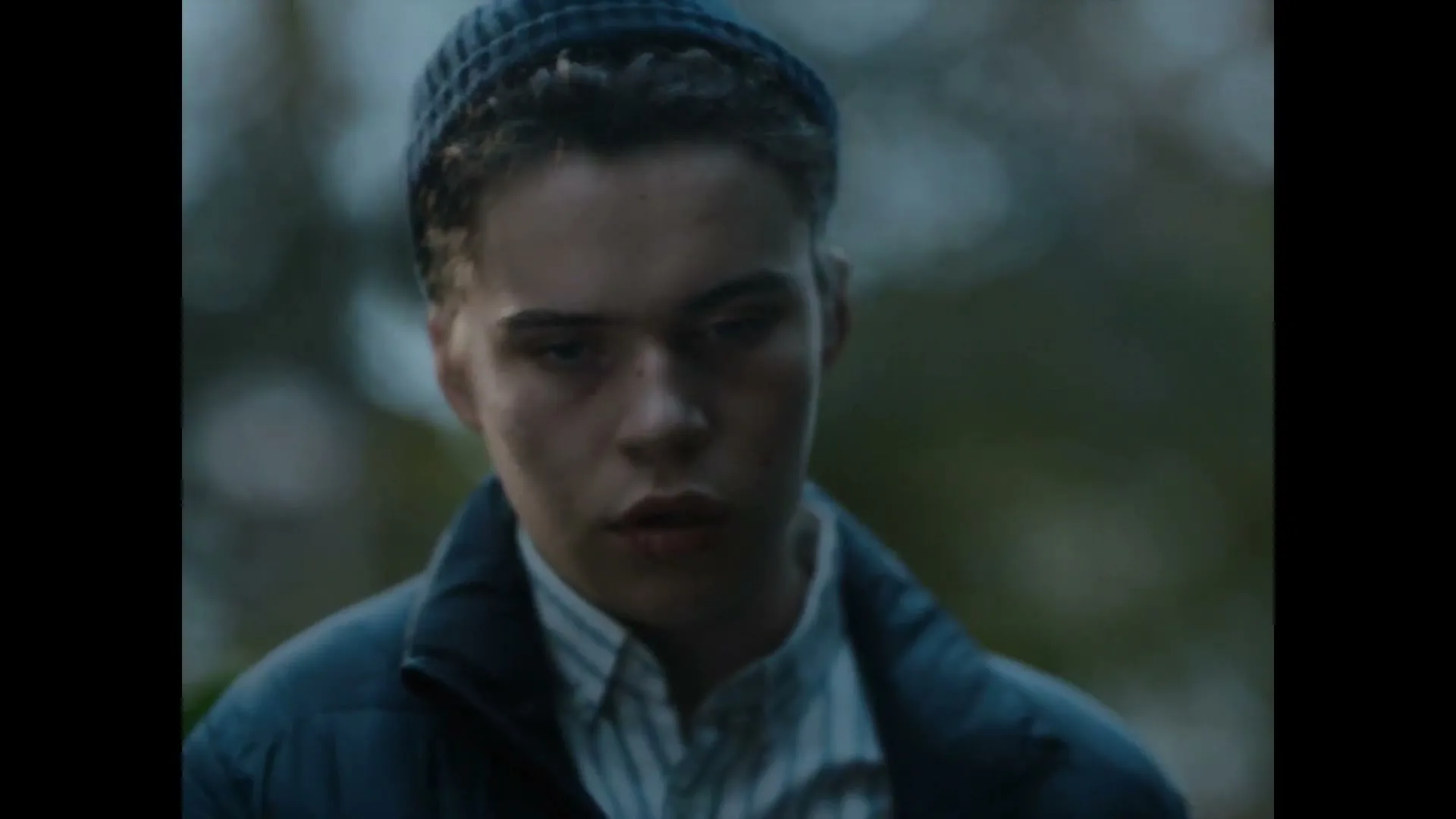 A young man with a blue beanie and jacket hugs an older woman, then looks directly at the camera with a serious expression in an outdoor setting.