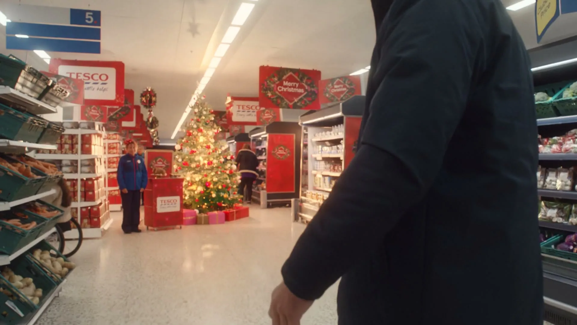 A person in a dark coat walks through a brightly lit supermarket aisle, past a decorated Christmas tree and festive displays.