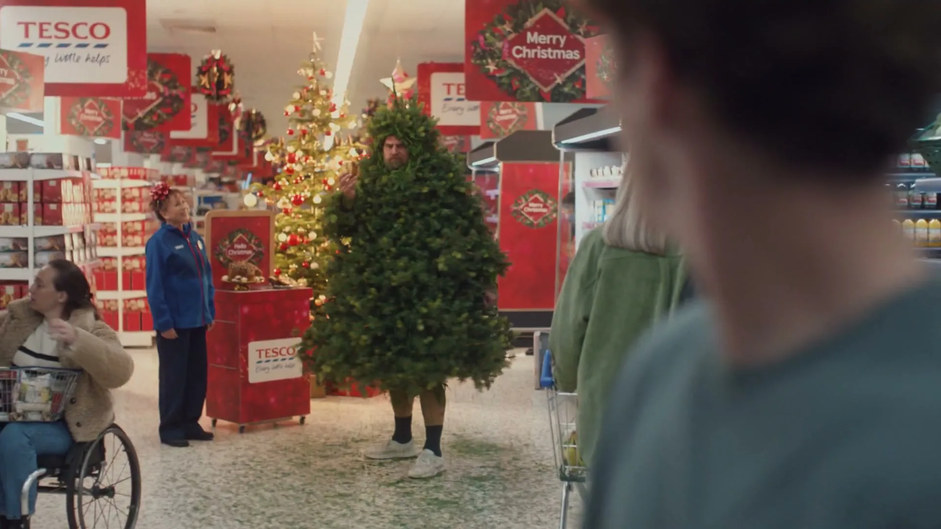 A man dressed as a Christmas tree stands in a supermarket aisle, while a female employee and a woman in a wheelchair look on, surrounded by festive decorations.