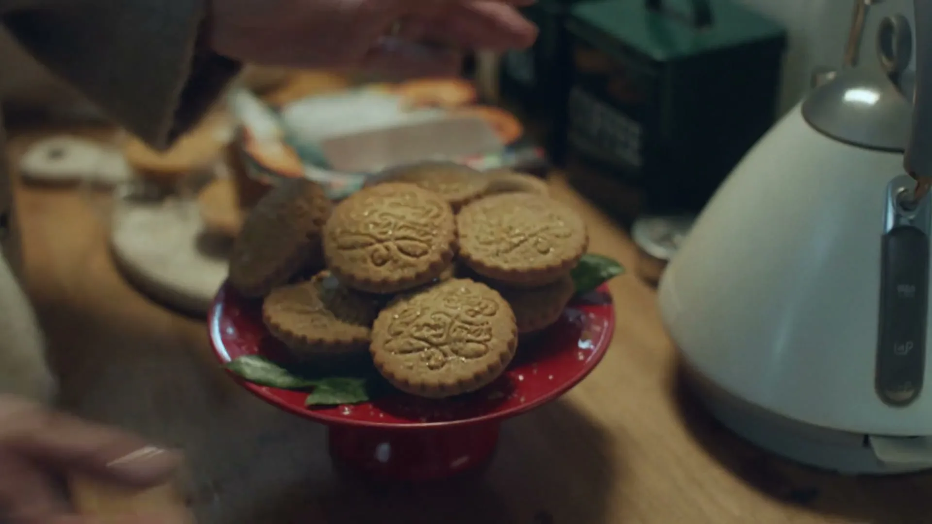 A person's hands arrange mince pies from a box onto a red cake stand on a wooden kitchen counter, next to a kettle and tea/coffee caddies.