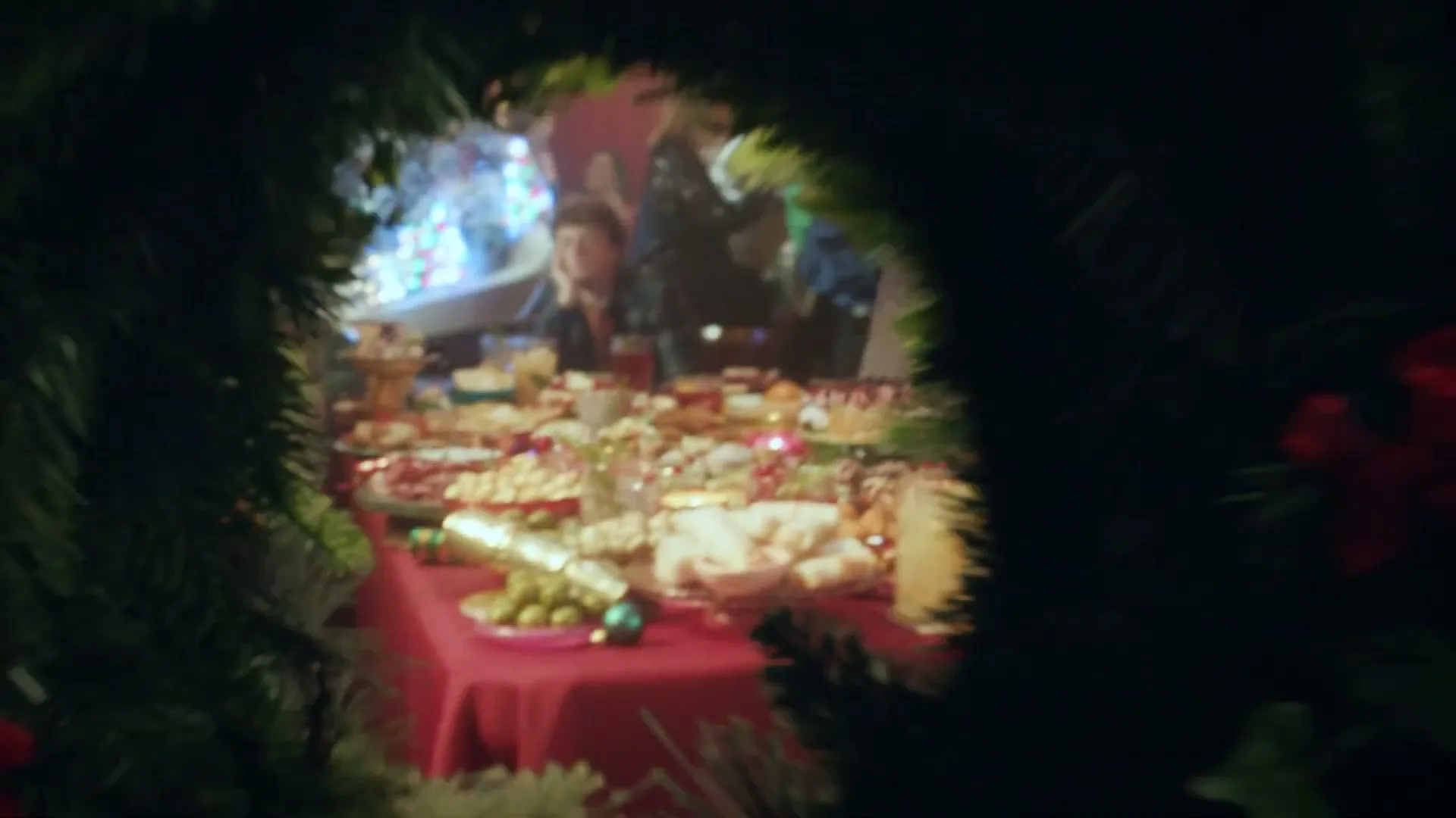 A camera moves through a lively Christmas party, passing by people in festive costumes and revealing a table laden with various holiday foods and drinks.