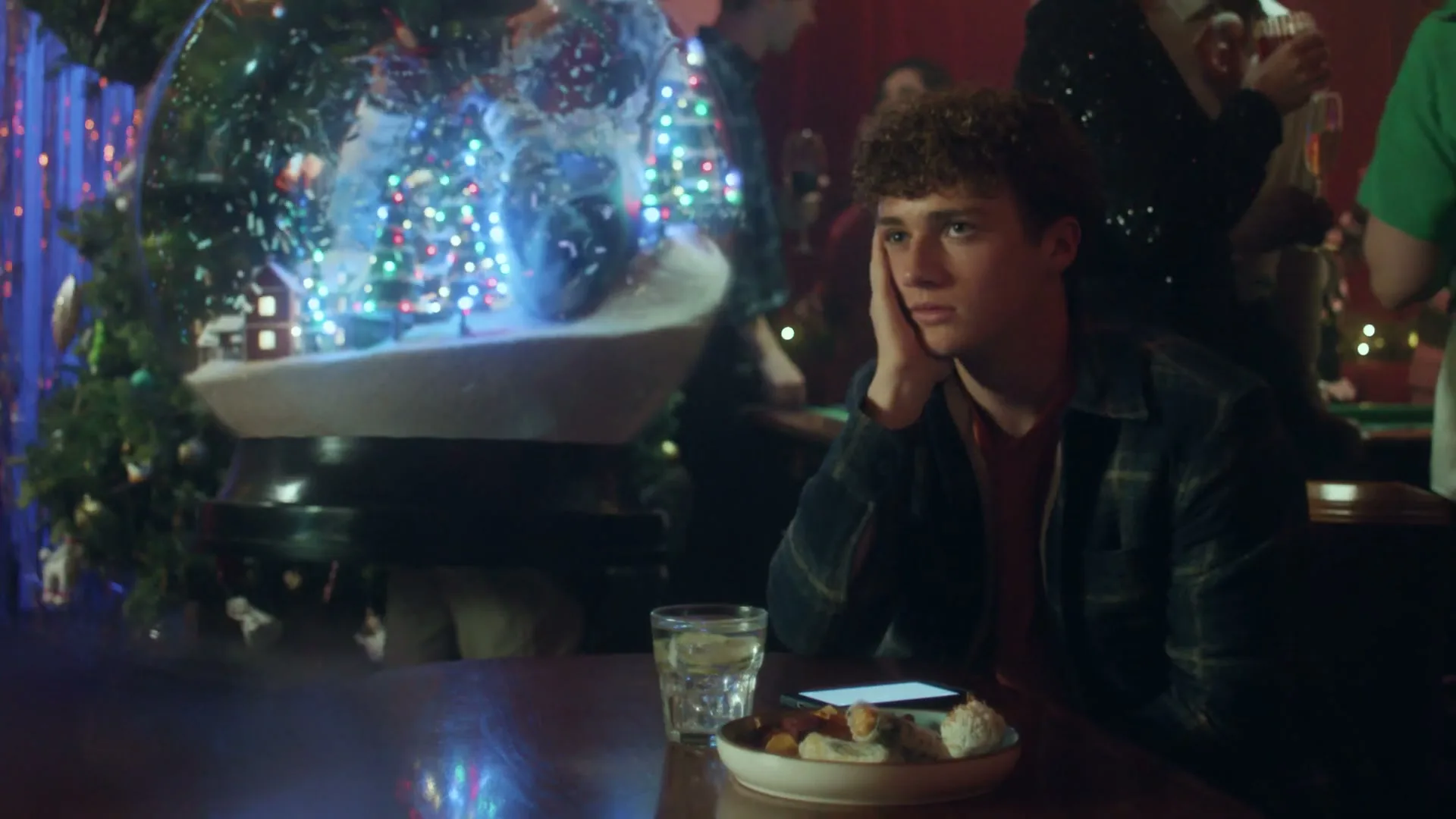 A young man with curly hair sits alone at a table, looking bored and resting his head on his hand, at a Christmas party.