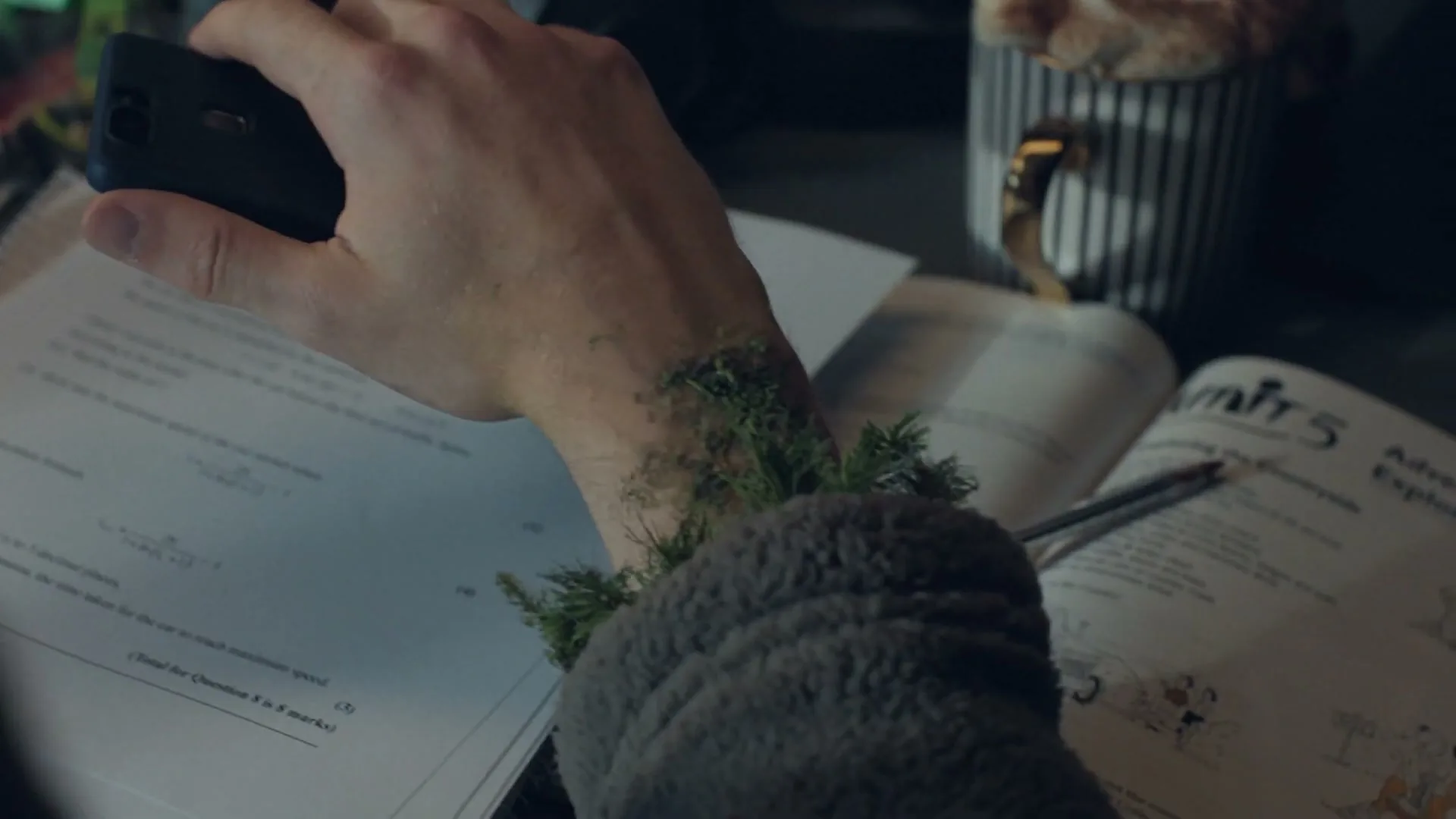 A person's arm, covered in green moss and pine needles, rests on a desk with open textbooks and a phone in hand, as if taking a photo.