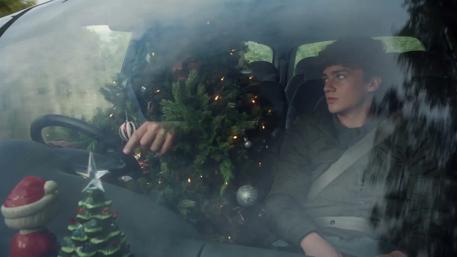 A man covered in Christmas tree branches and ornaments drives a car, while a young man in the passenger seat looks out the window.