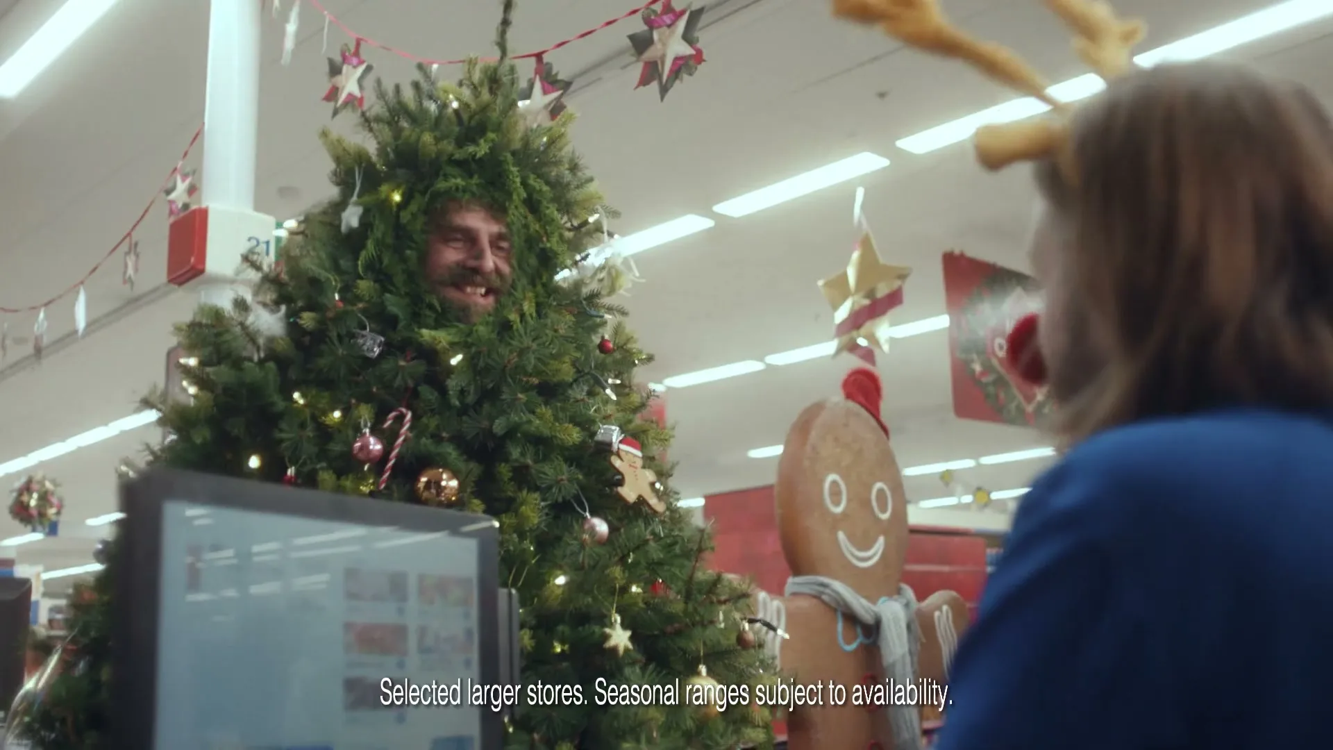 A man dressed as a Christmas tree with lights and ornaments smiles at a woman wearing reindeer antlers in a brightly lit supermarket.