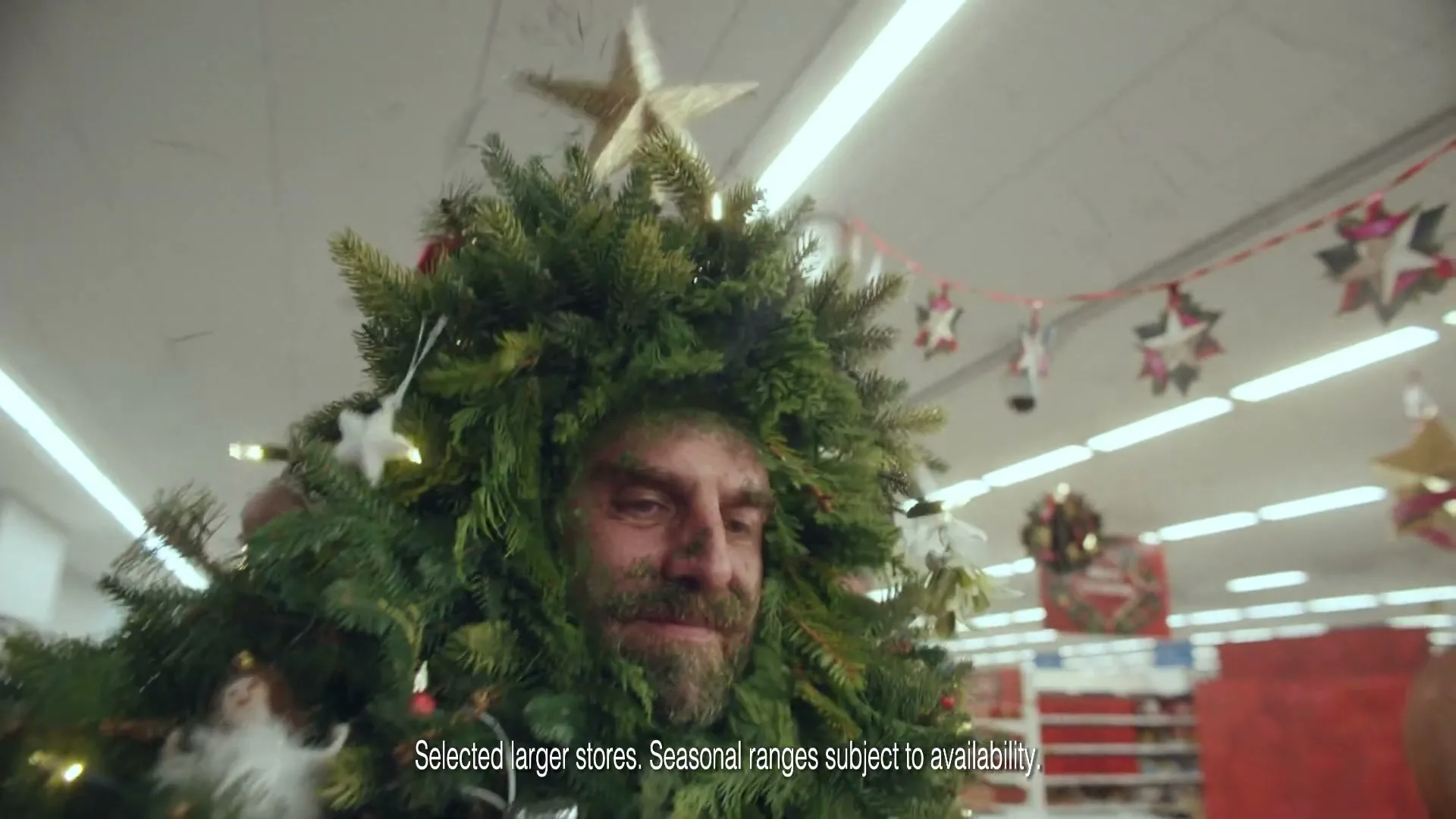 A man dressed as a Christmas tree with green face paint and a gold star on his head looks around a supermarket aisle.