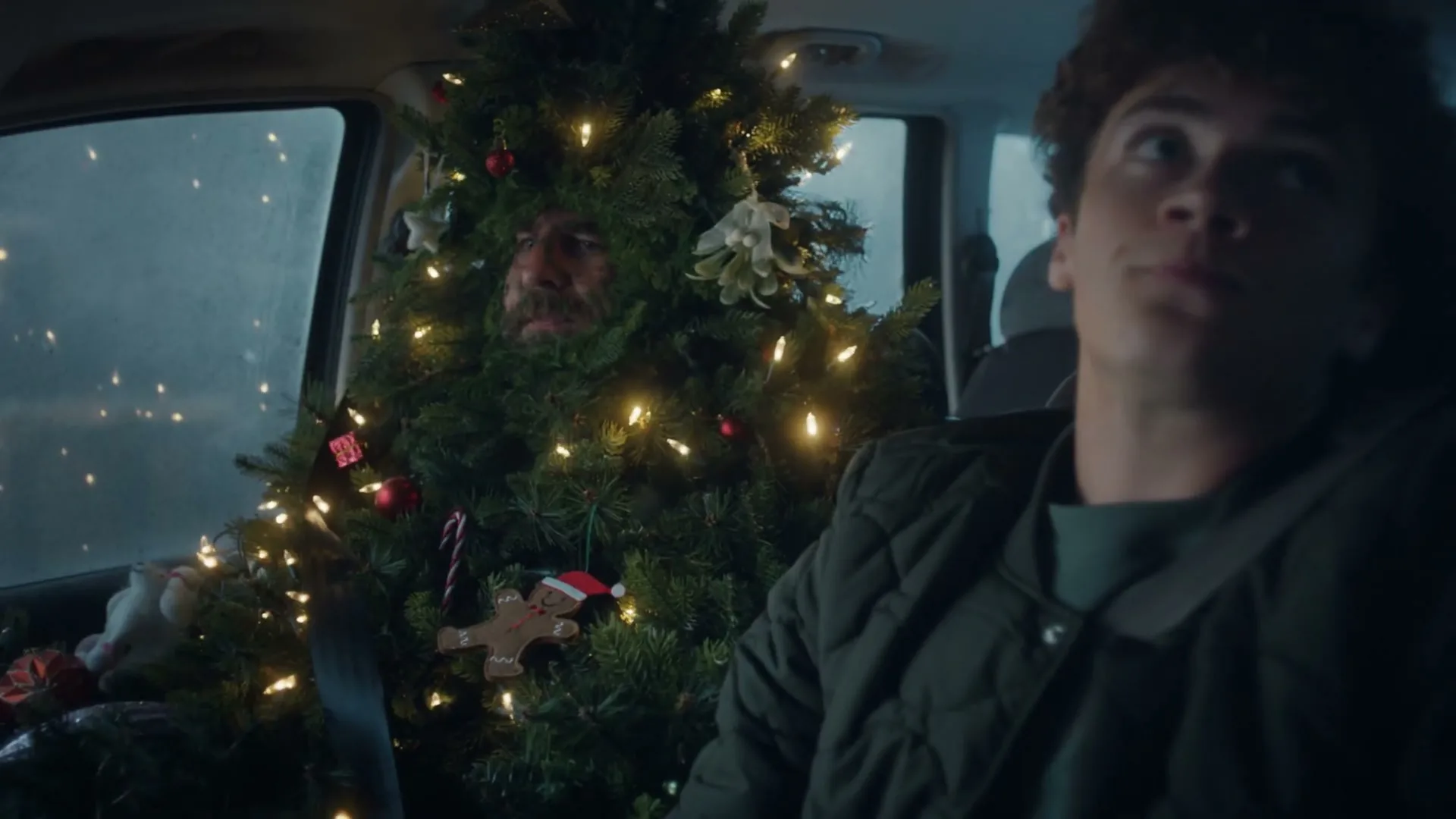 A young man in a car looks forward, while a man dressed as a Christmas tree sits in the back seat, decorated with lights and ornaments.