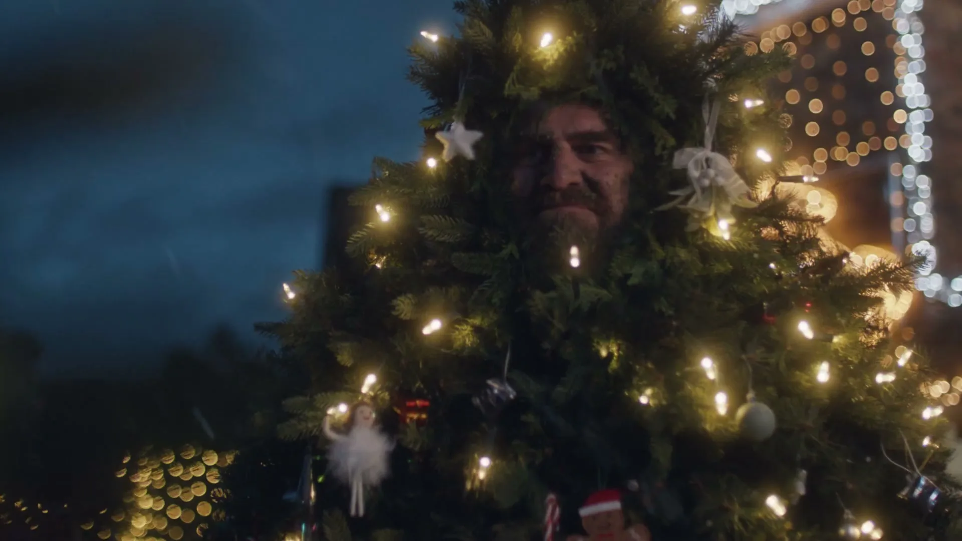 A man with a beard and dirty face is dressed as a Christmas tree, adorned with lights and ornaments, standing outside at night.