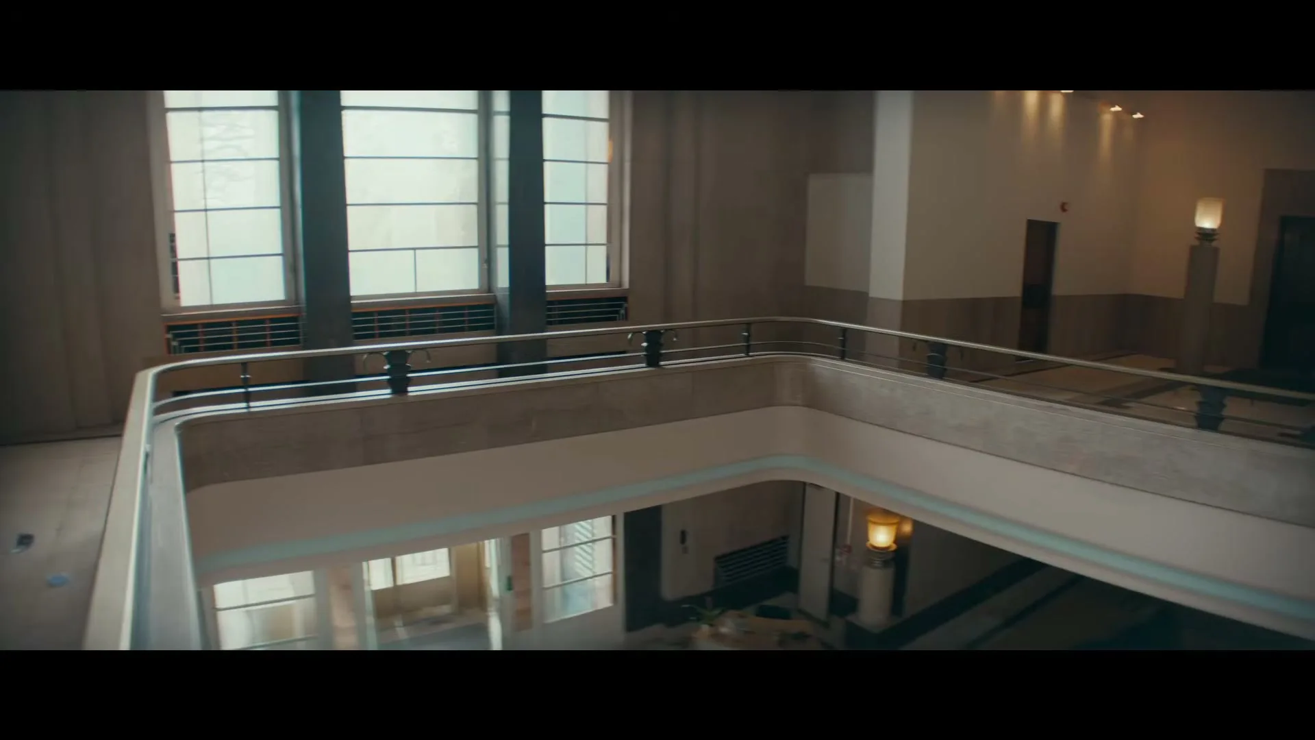Three masked figures in hoodies and carrying bags run through a grand, empty building, descending a staircase and crossing a large hall.