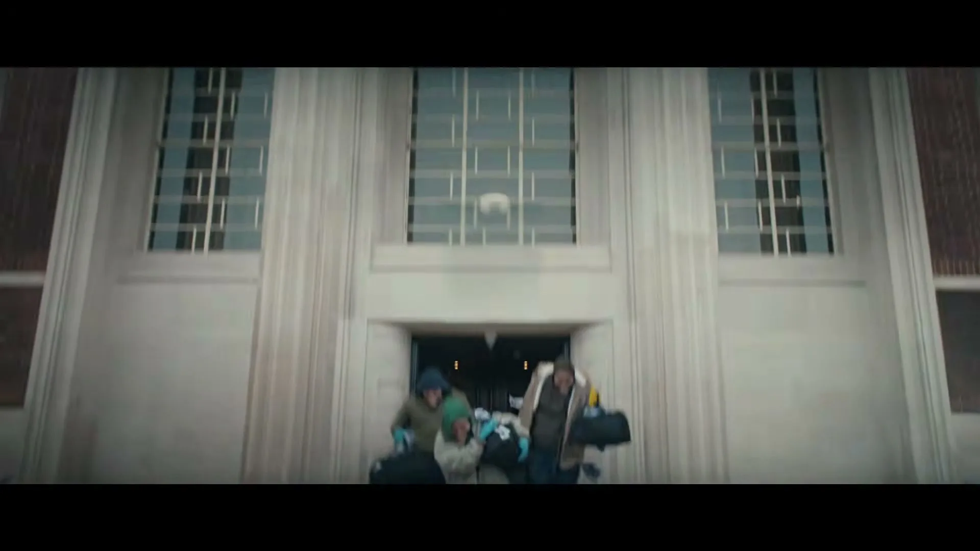 Three masked bank robbers, two men and one woman, run out of a bank carrying bags of money, looking happy and excited.