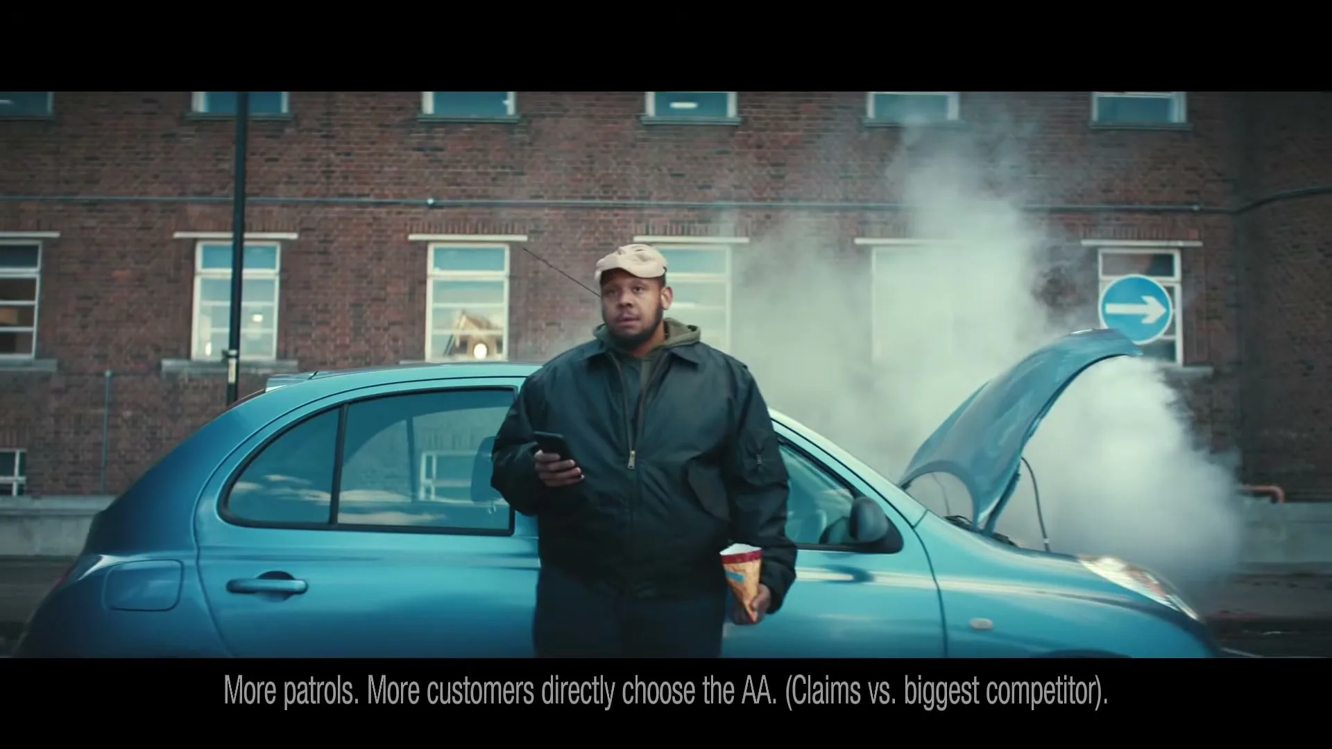 A man in a cap and jacket leans against a broken-down blue car with smoke coming from the engine, holding a phone and a drink, on a city street.