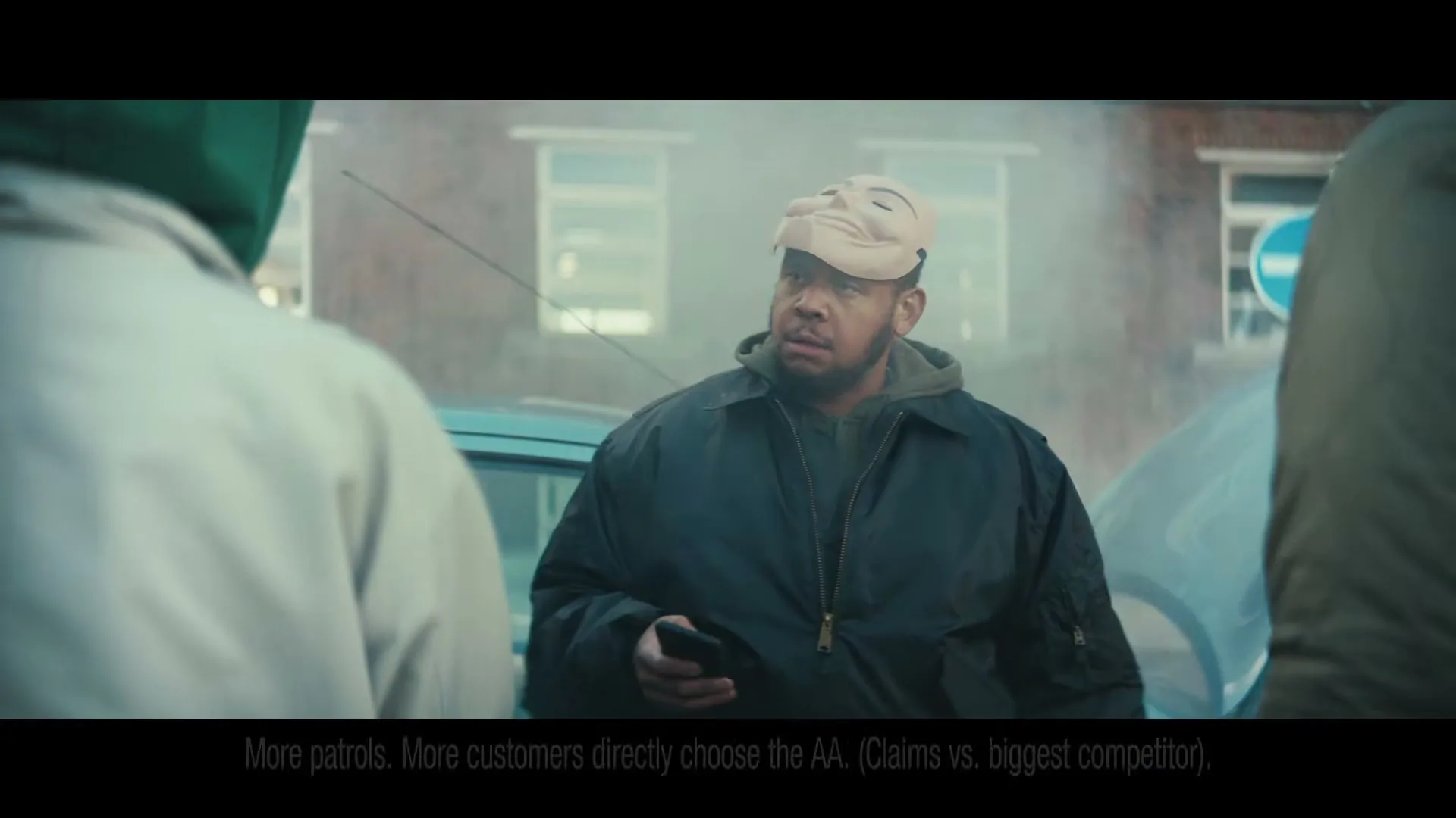 A man in a beige mask and dark jacket holds up a phone to two other men in a misty urban street.