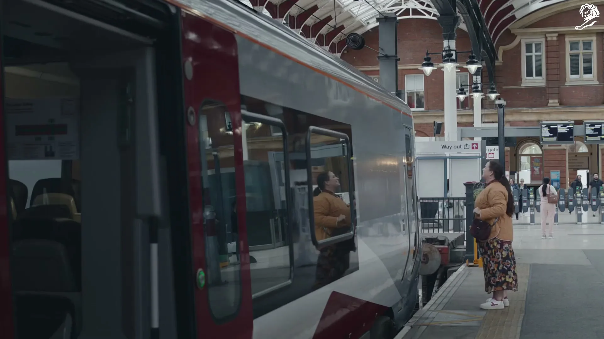A woman in a yellow jacket and floral skirt stands on a train platform, looking into an open train door.