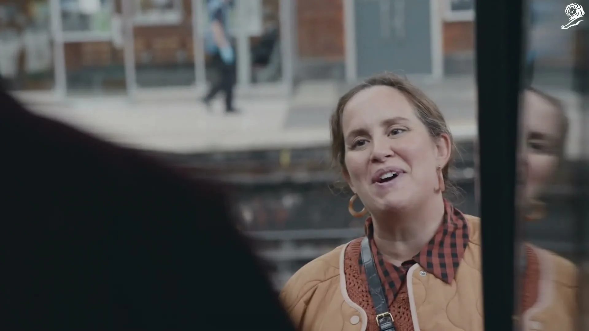 A woman with brown hair and a quilted jacket smiles and talks to someone off-screen, with a blurry street scene in the background.