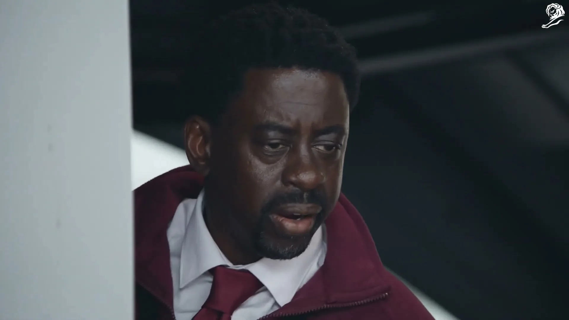 A Black man in his 40s, wearing a white shirt, red tie, and maroon jacket, looks around with a concerned expression inside a vehicle.