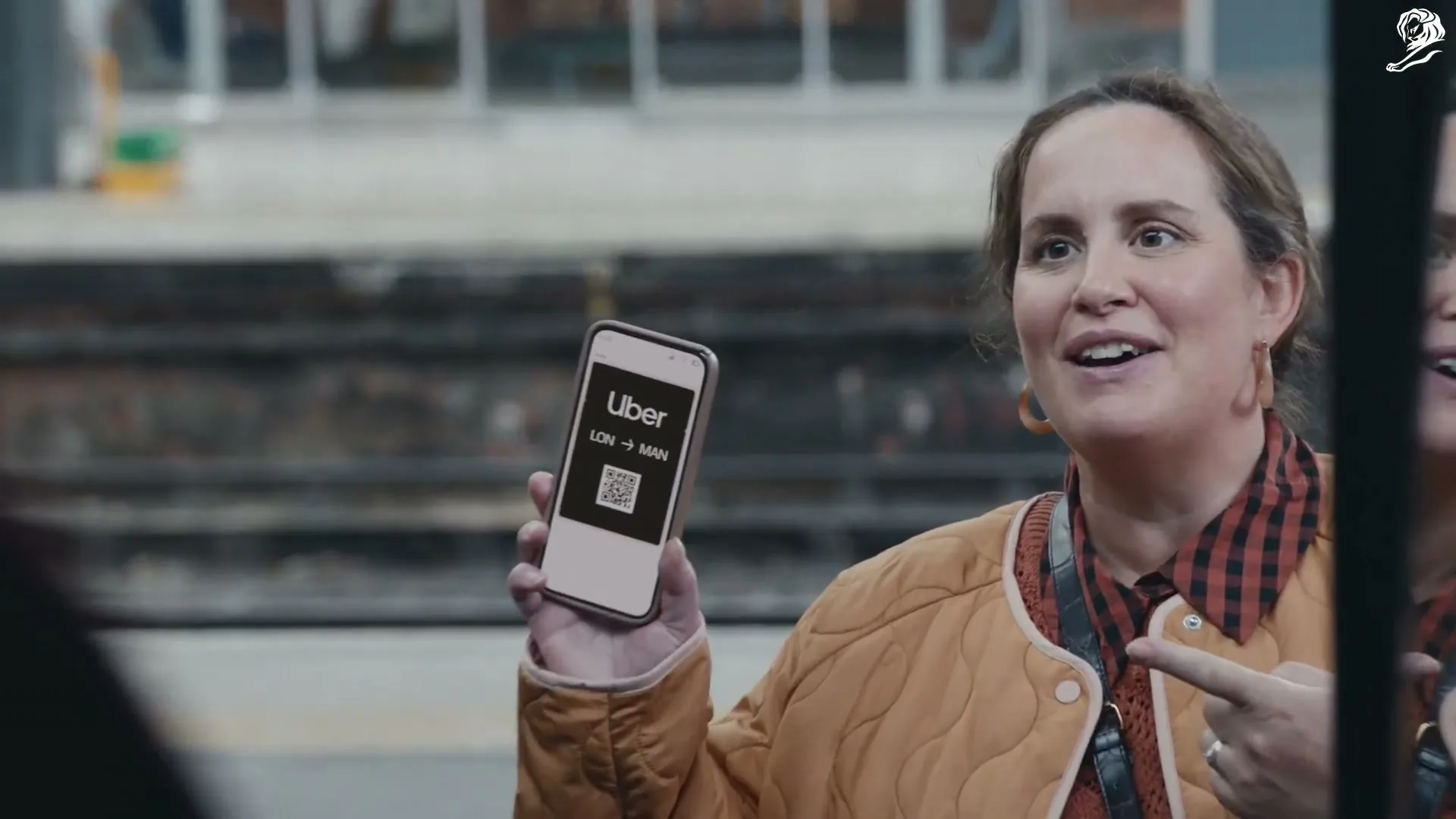 A woman in a quilted jacket and plaid shirt smiles, holding up her phone with the Uber app open, pointing to it, at a train station.