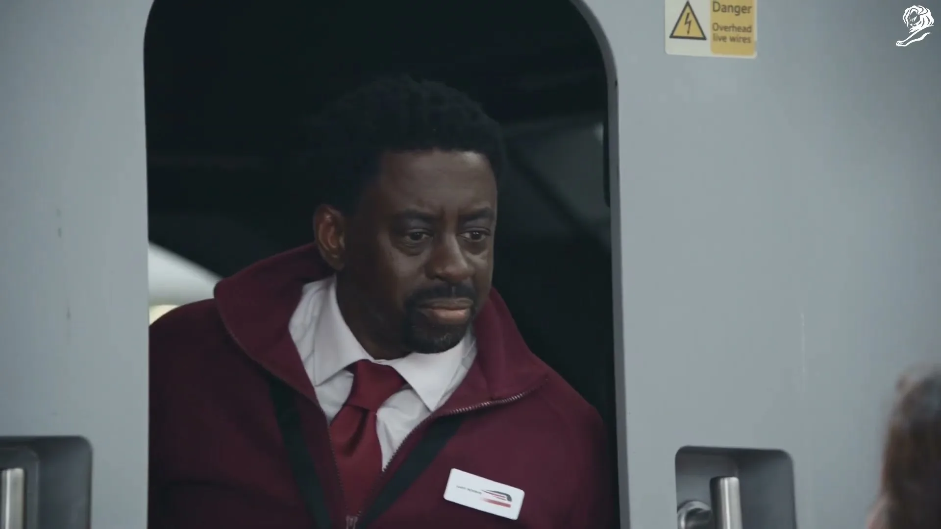 A male train conductor in a maroon jacket and red tie looks surprised and then smiles at someone off-camera inside a train.