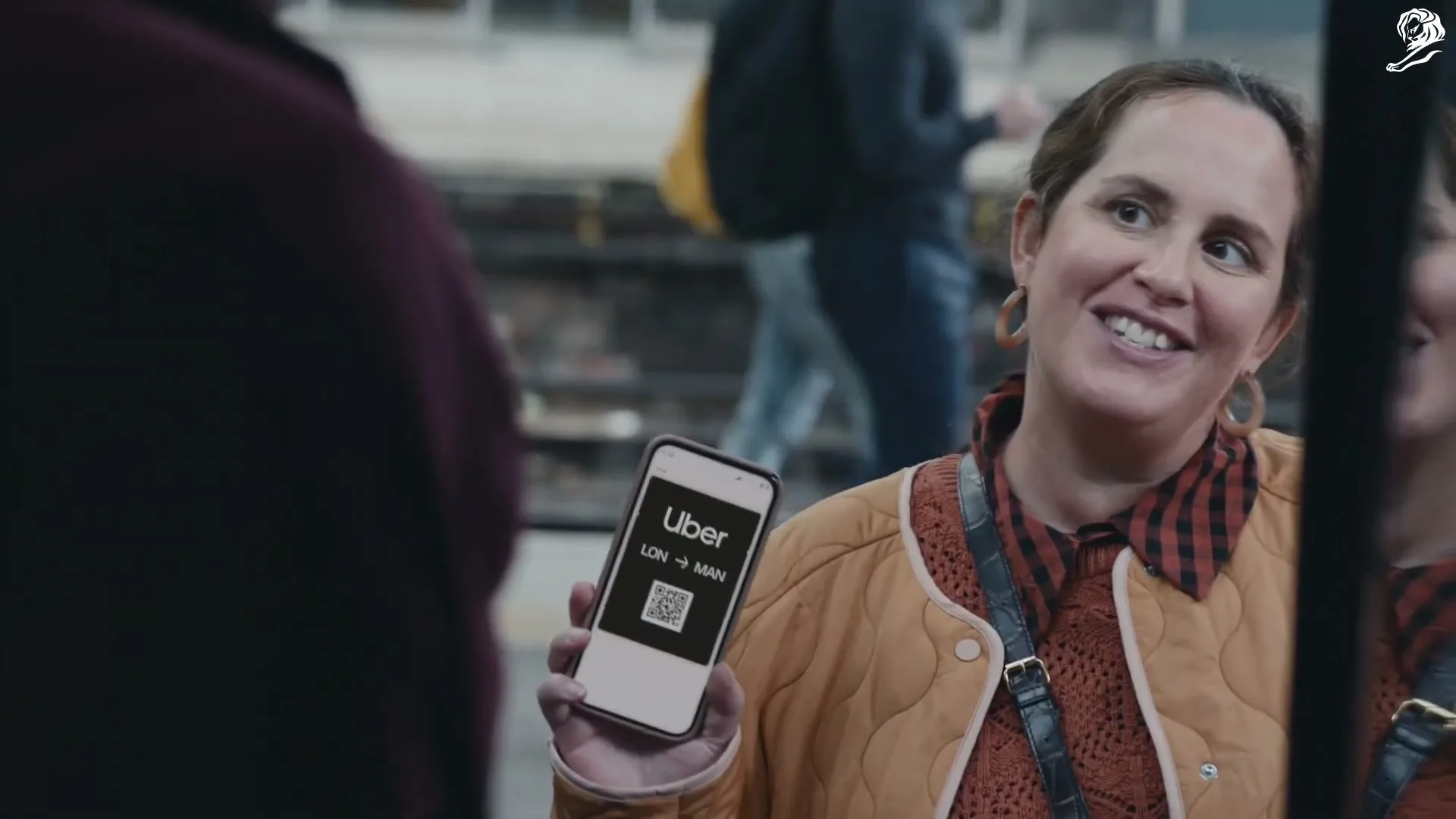 A woman in a quilted jacket holds up her phone with the Uber app open, showing a QR code and "LON -> MAN" on screen, as she talks to someone off-screen.