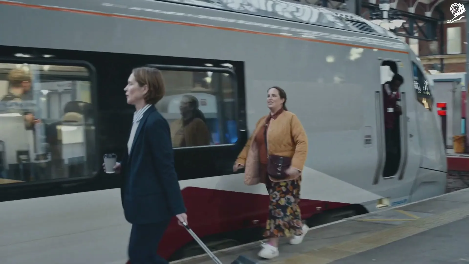 A woman in a quilted jacket and patterned skirt smiles and holds up her phone with the Uber app open, then rushes to catch a train at a station.