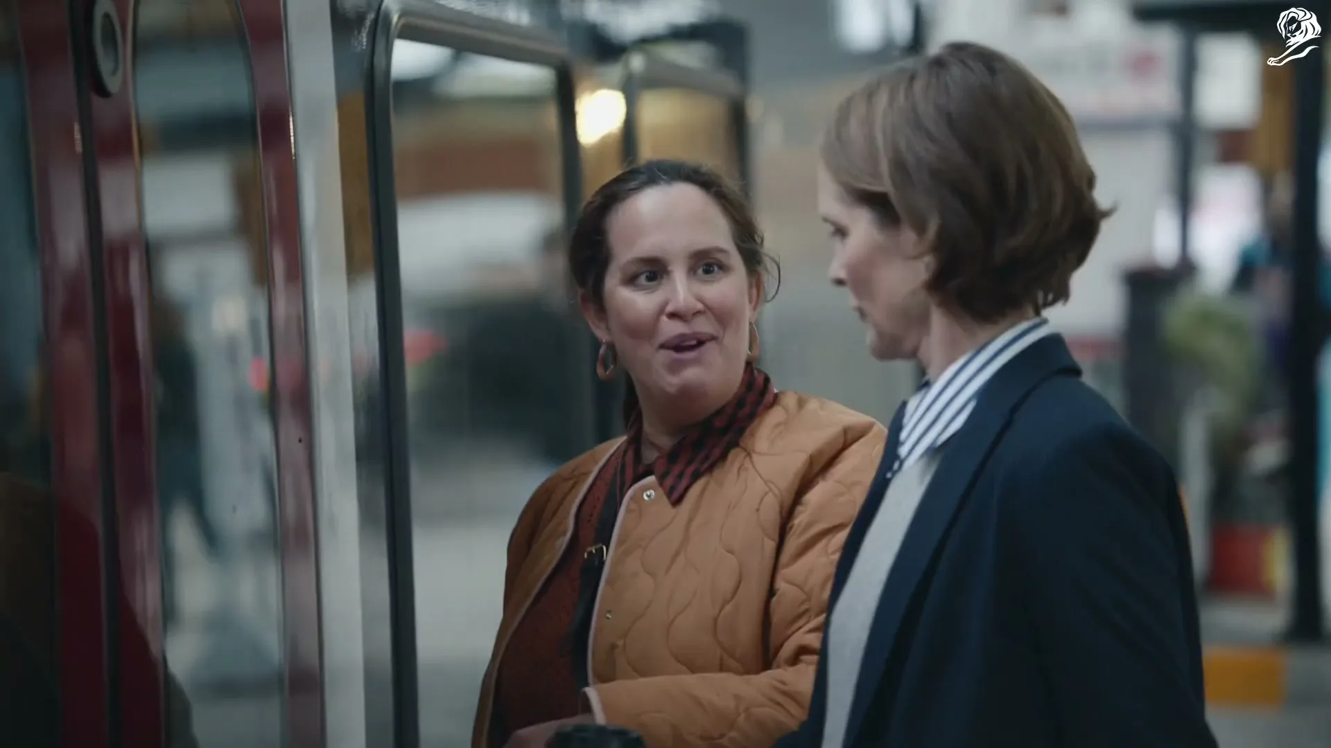 Two women, one in a quilted jacket and the other in a blazer, stand next to a vehicle, talking and smiling.