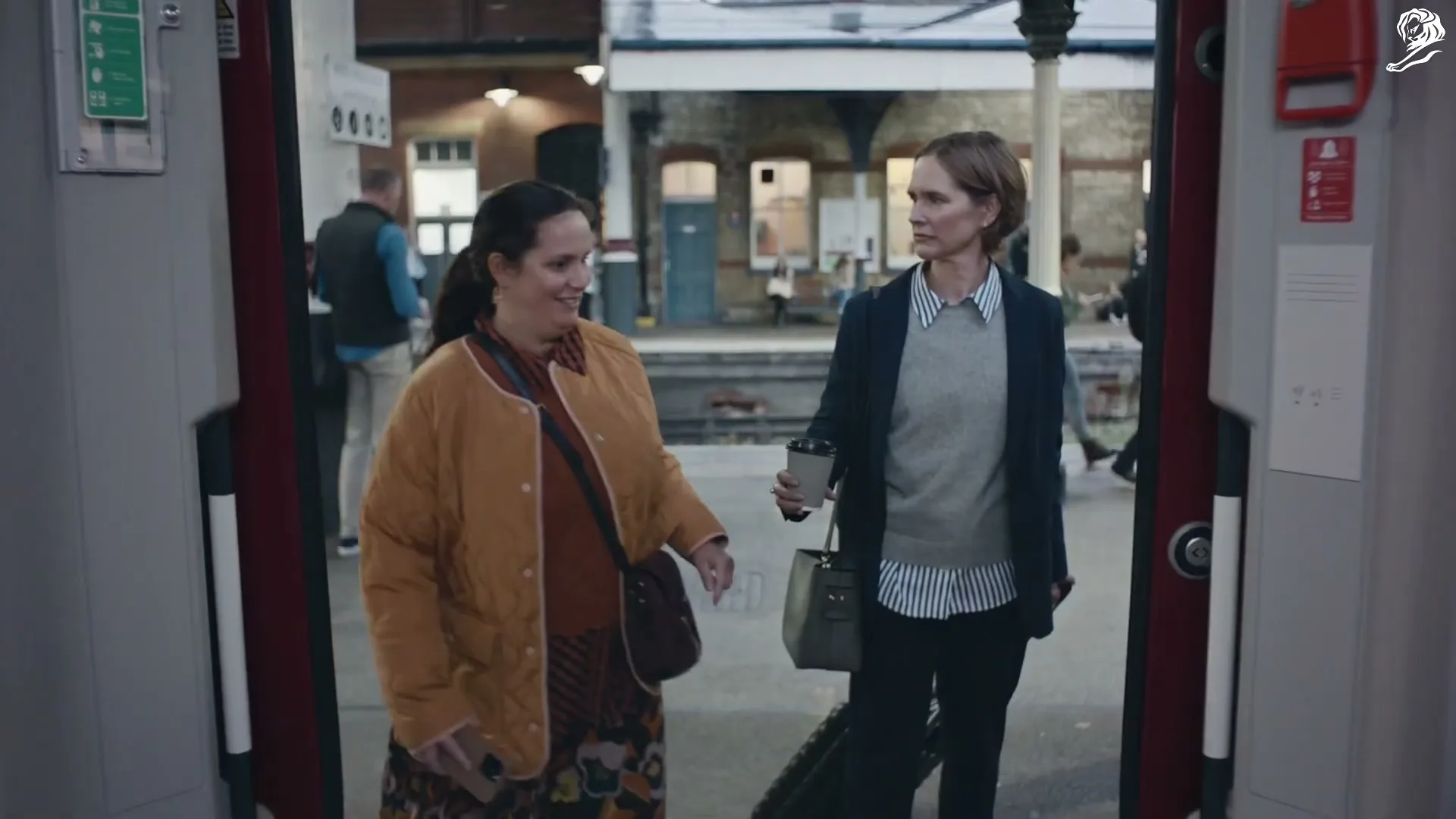 Two women, one in a brown jacket and the other in a grey sweater and black blazer, exit a train onto a platform at a train station.
