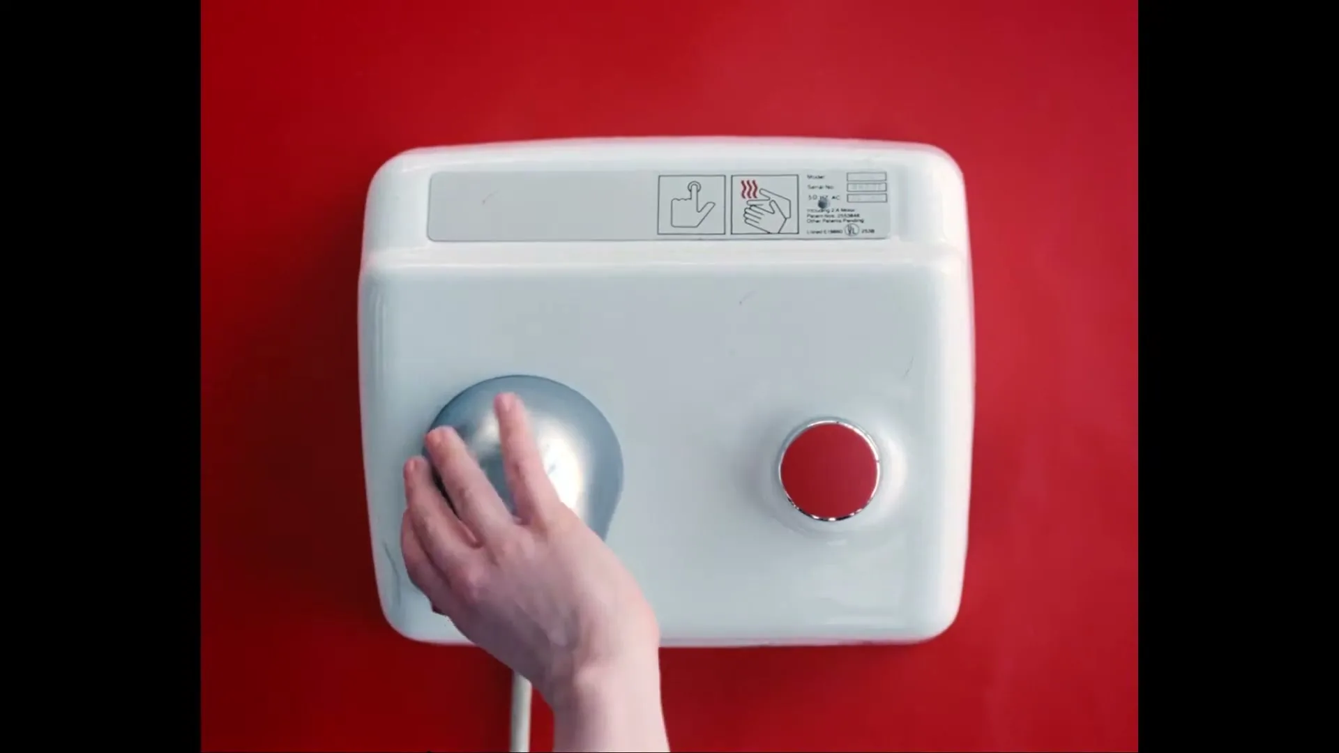 A white hand dryer with a red button is mounted on a red wall, as a person's hand moves to activate it.