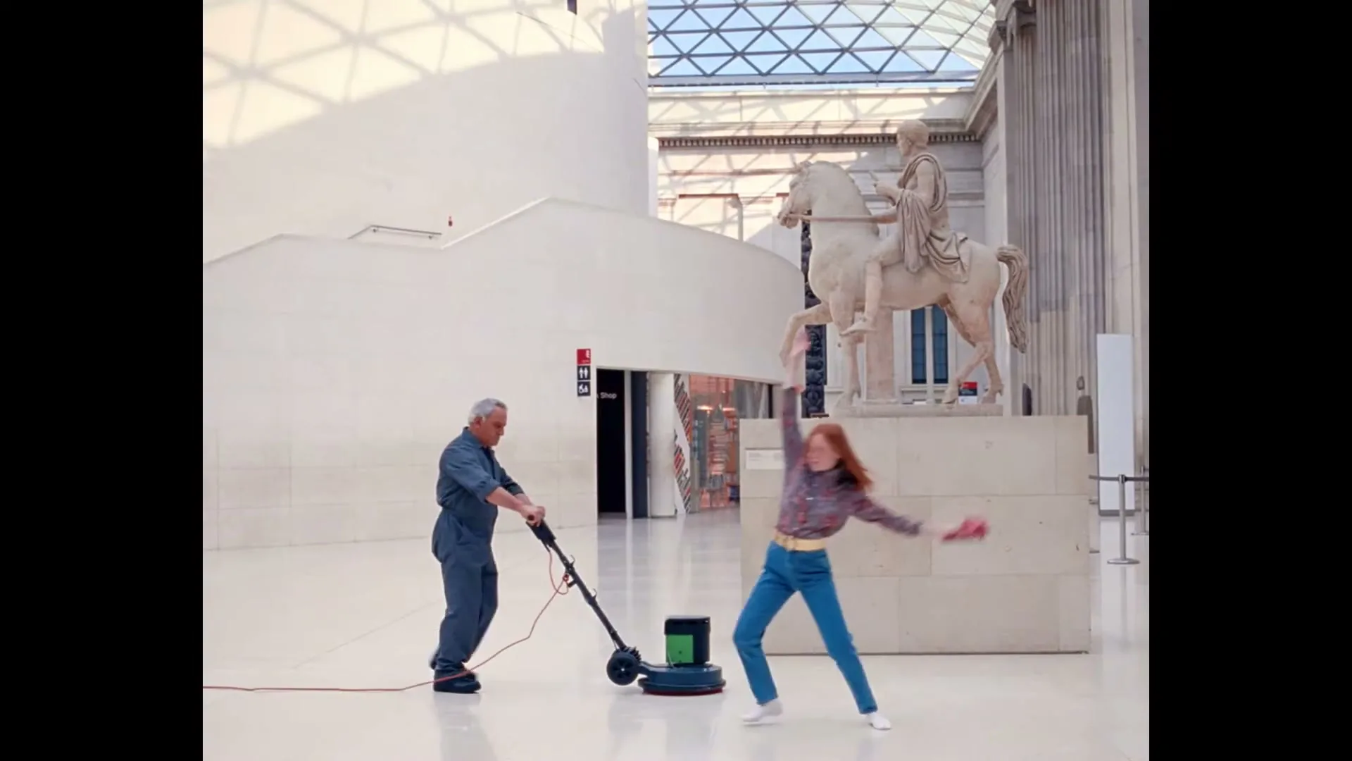 A janitor in blue overalls polishes a shiny white floor with a machine, while a young woman with red hair dances past him in a museum.
